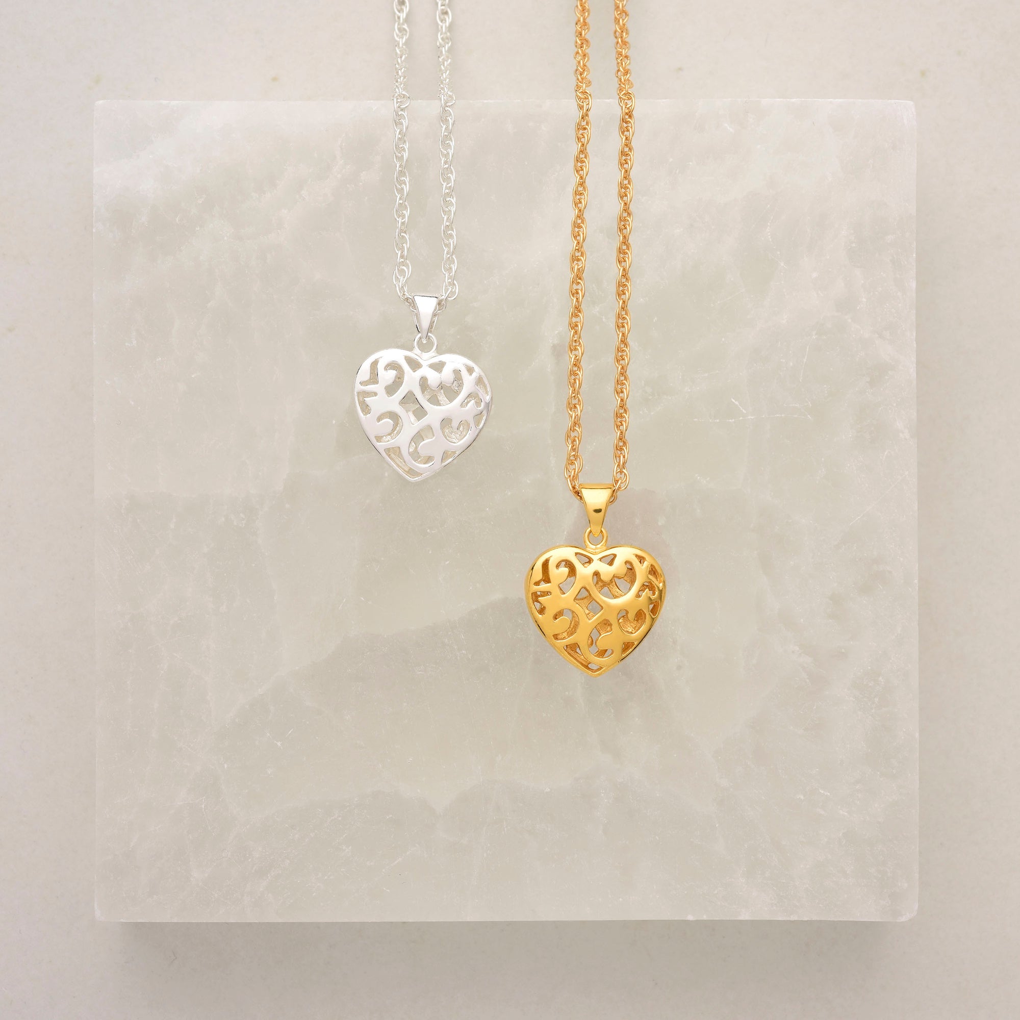 Two heart-shaped necklaces with ornate cut-out designs, one silver on a silver chain and one gold on a gold chain, displayed on a translucent, light stone background.
