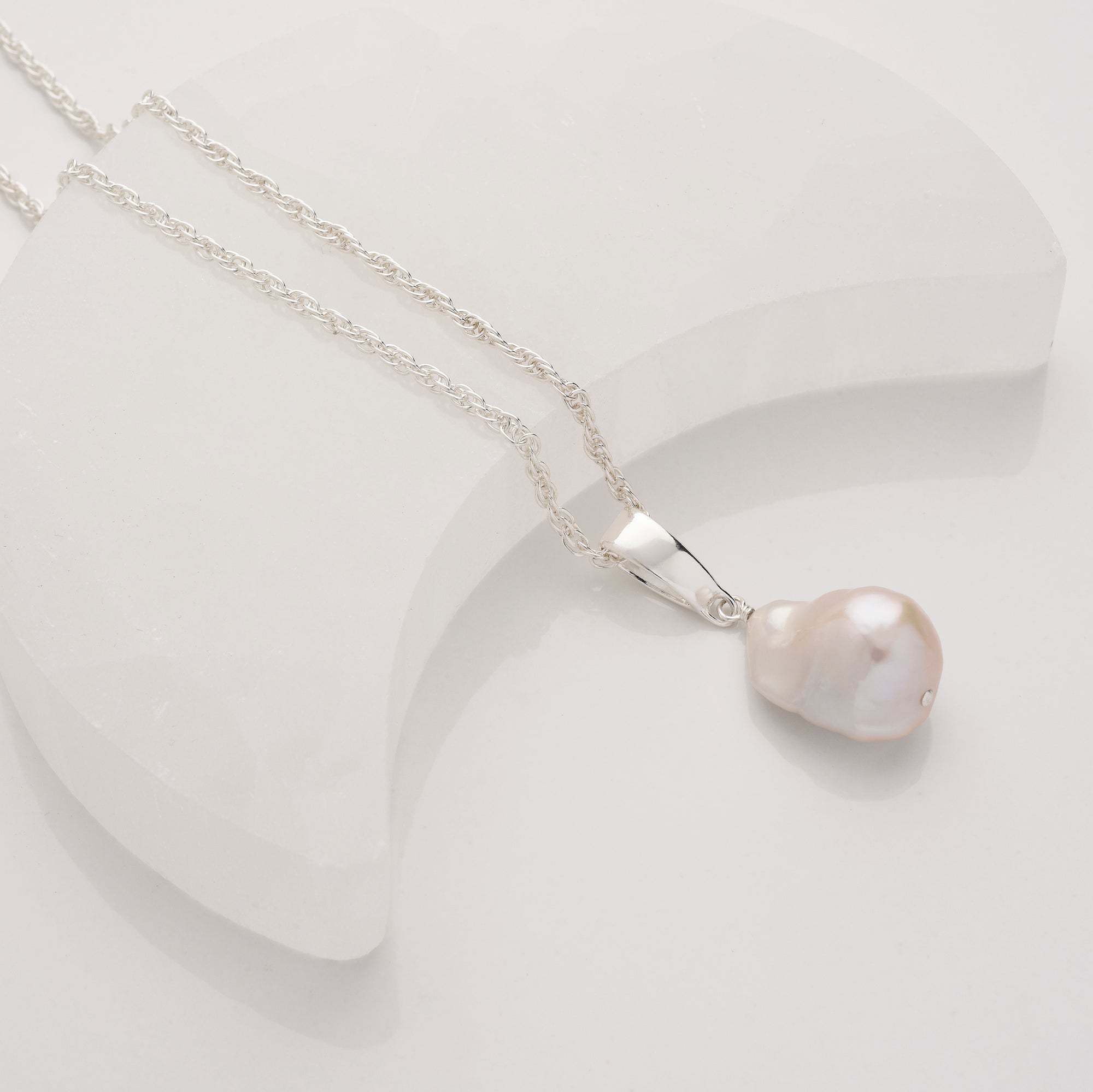 A silver chain necklace with a large, irregular-shaped white pearl pendant, displayed on a smooth, white, crescent-shaped surface.