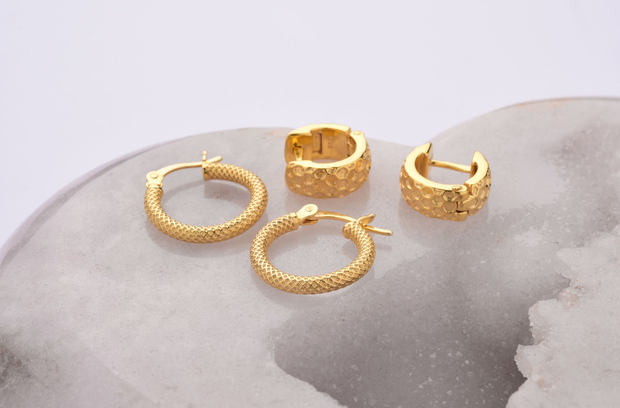 Two pairs of gold hoop earrings, one with a textured woven pattern and the other with a hammered finish, are displayed on a white and gray marble-like surface.