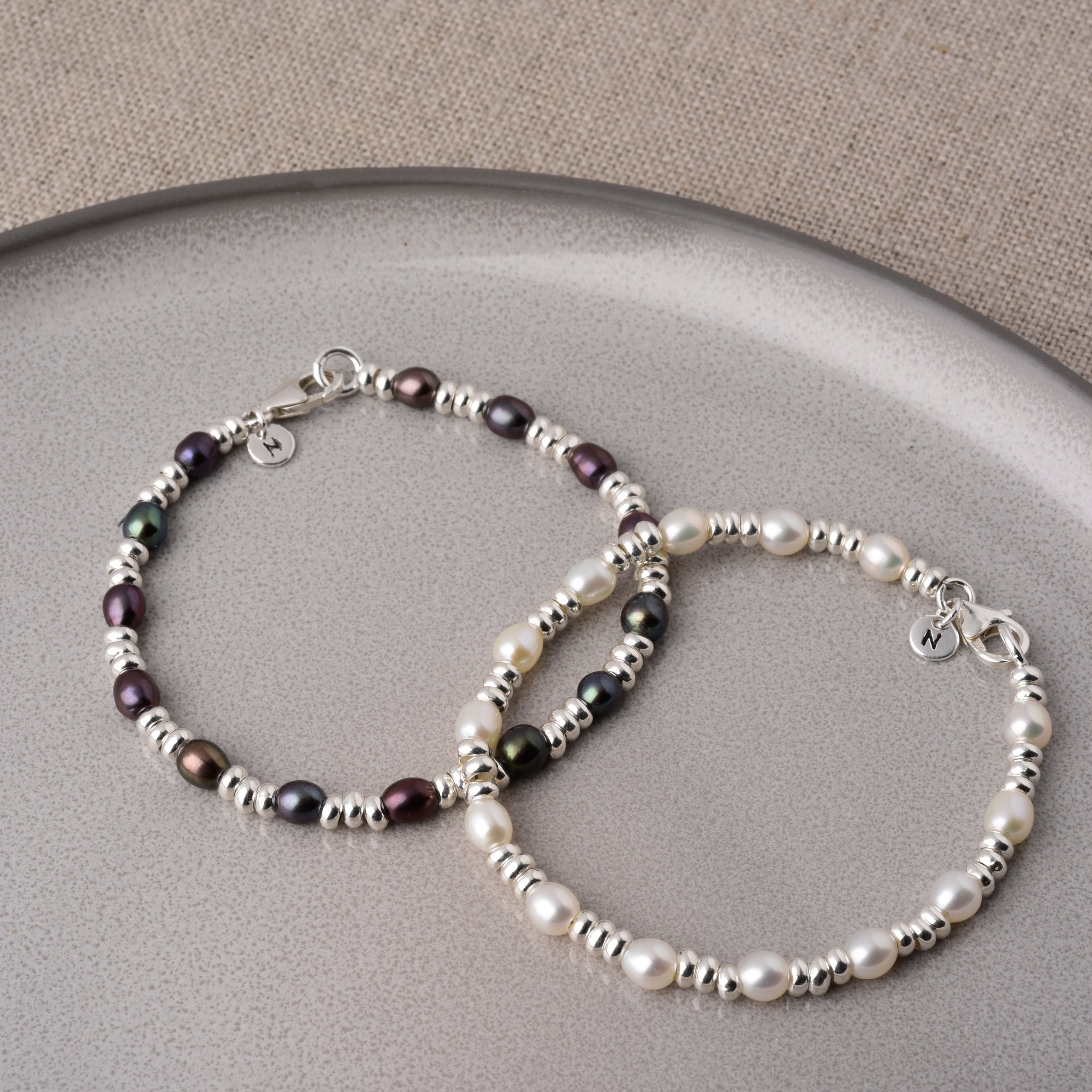 Two beaded bracelets with silver accents, one featuring dark pearls and the other white pearls, displayed on a curved, light gray dish with a textured beige background.