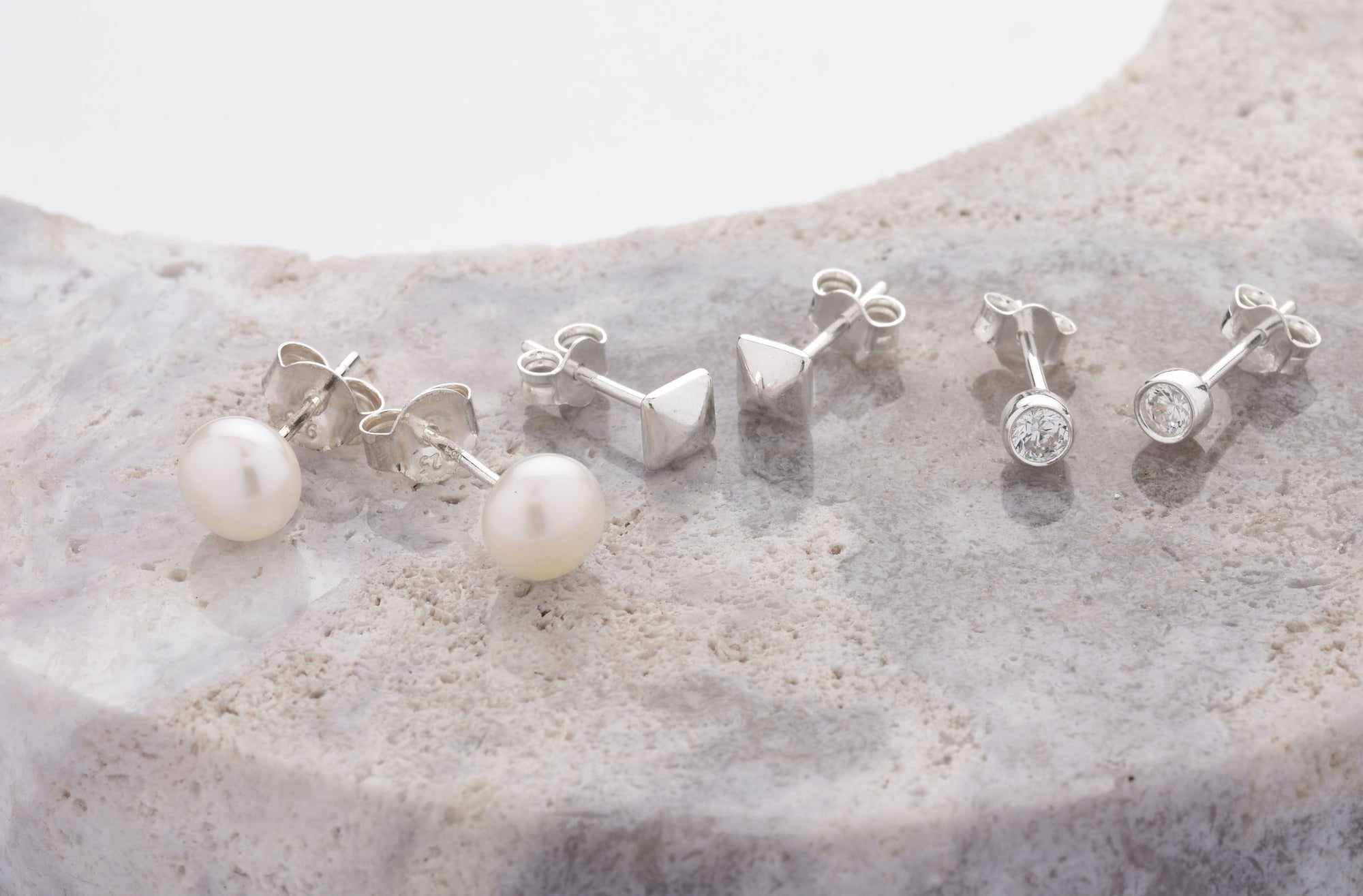 Three pairs of silver stud earrings on a stone surface: one with pearls, one with geometric metal shapes, and one with round clear gemstones.