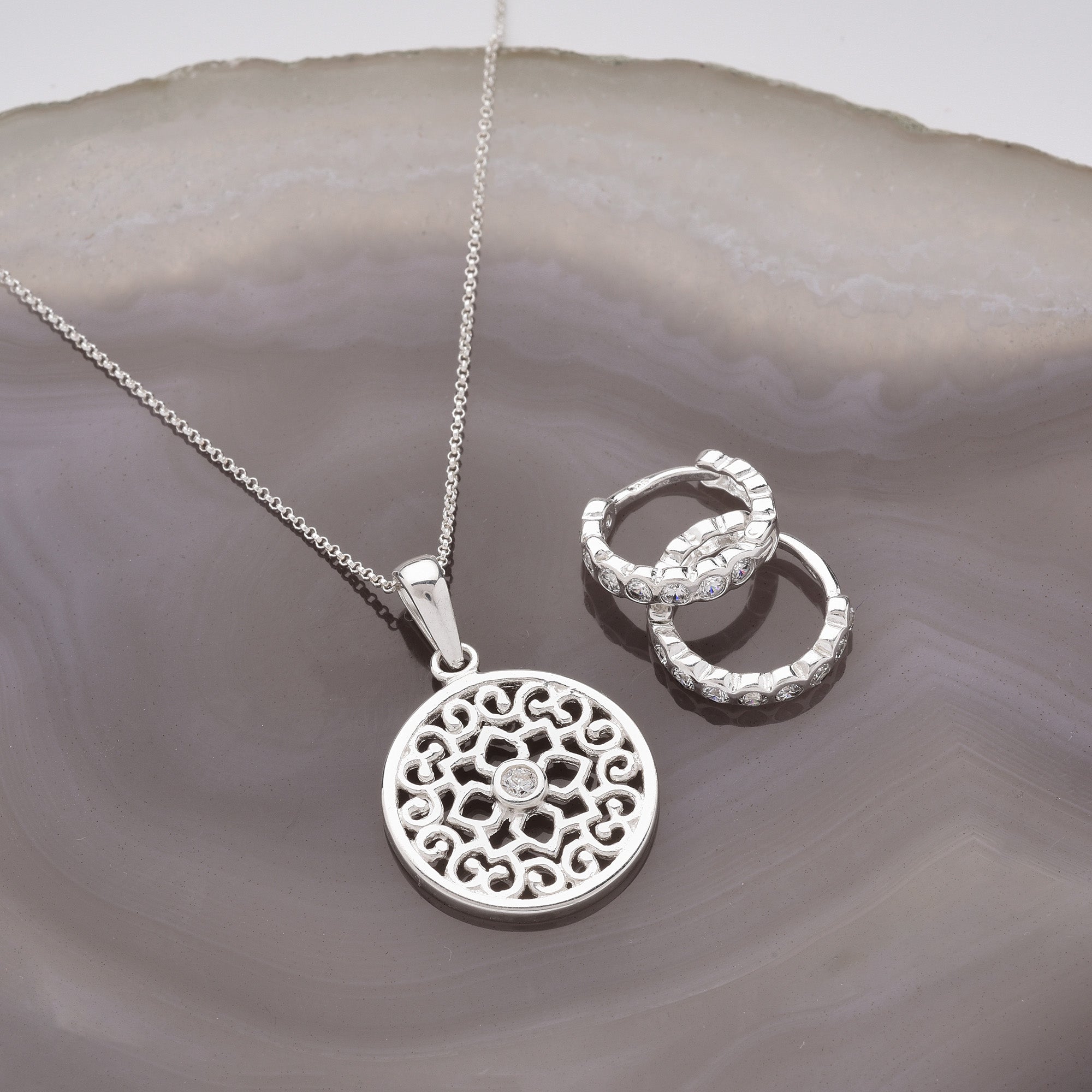 A silver necklace with an intricate round pendant and a pair of textured silver hoop earrings rest on a polished agate slice with shades of gray and white.