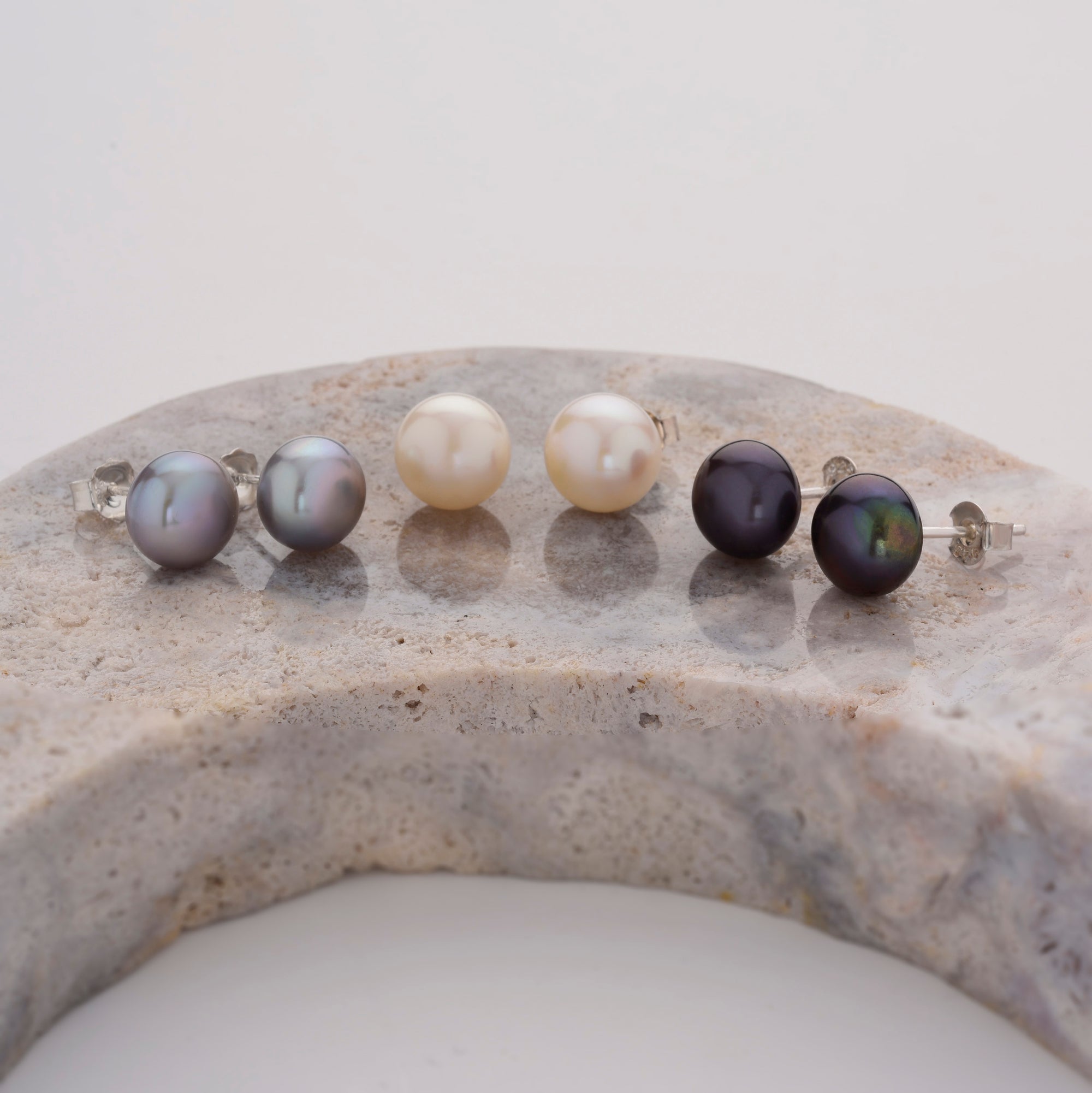 Three pairs of pearl stud earrings—gray, white, and black—are displayed in a row on a curved, light-colored stone surface against a plain background.