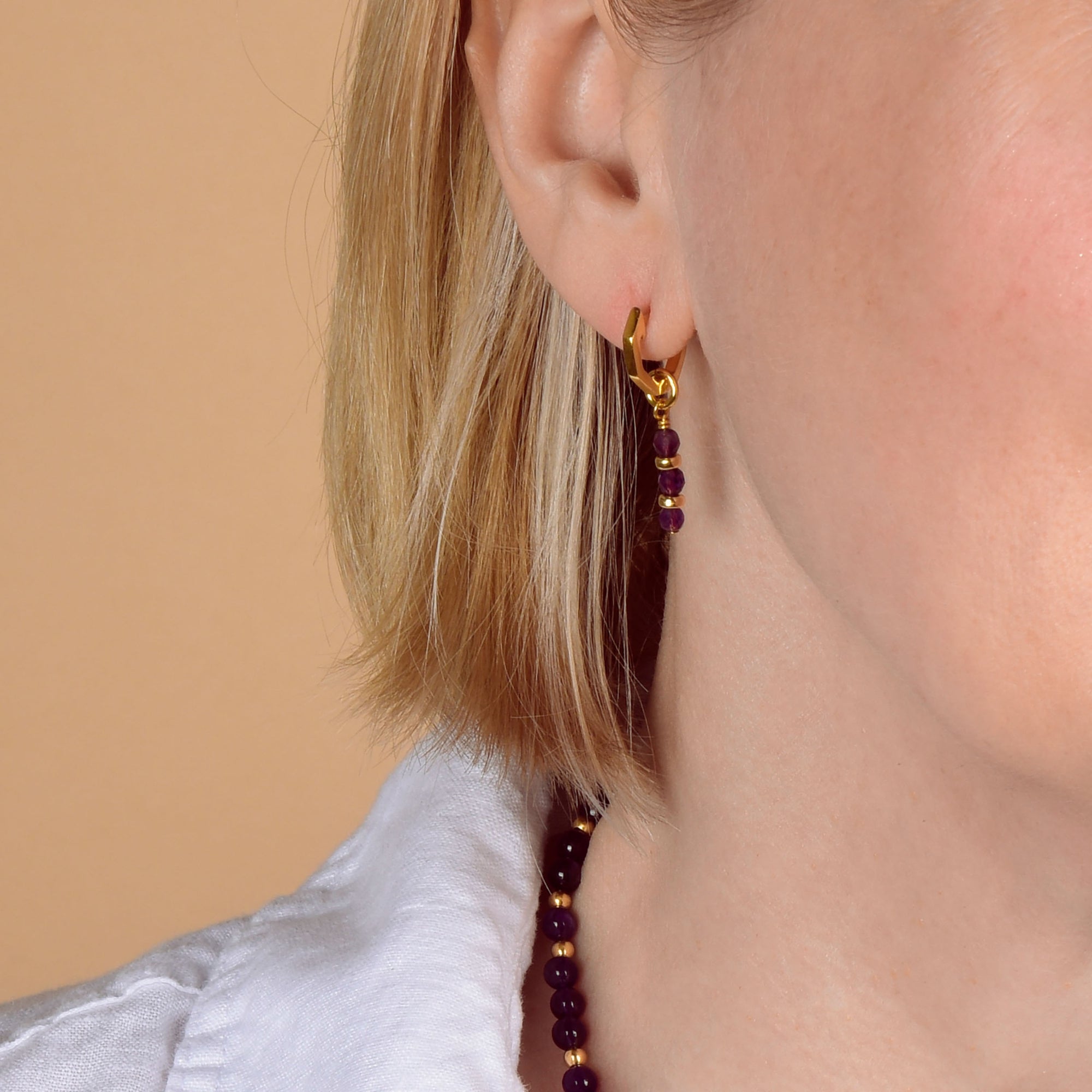 Close-up of a woman’s ear with short blonde hair, wearing the Garnet Linear Huggie Birthstone Earrings for January—delicate gold hoops adorned with rich garnet beads. A matching beaded necklace and the collar of a white shirt are also visible, set against a soft beige background.