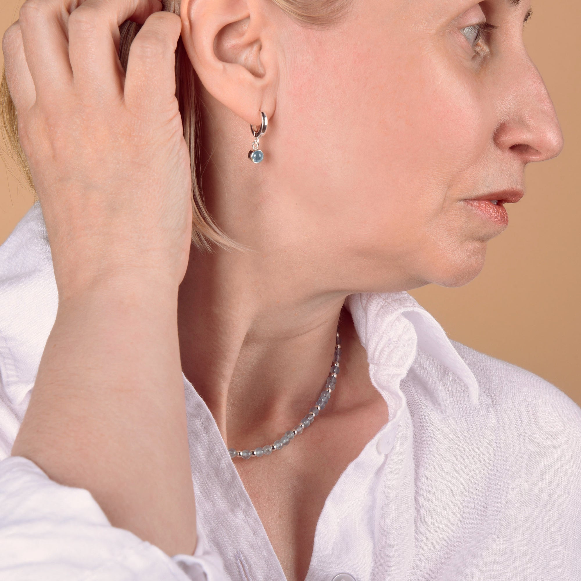 A woman in a white shirt touches her ear, showcasing the Aquamarine March Gem Hoop Birthstone Earrings—sterling silver hoops adorned with blue aquamarine stones—paired with a matching beaded necklace, set against a peach background.