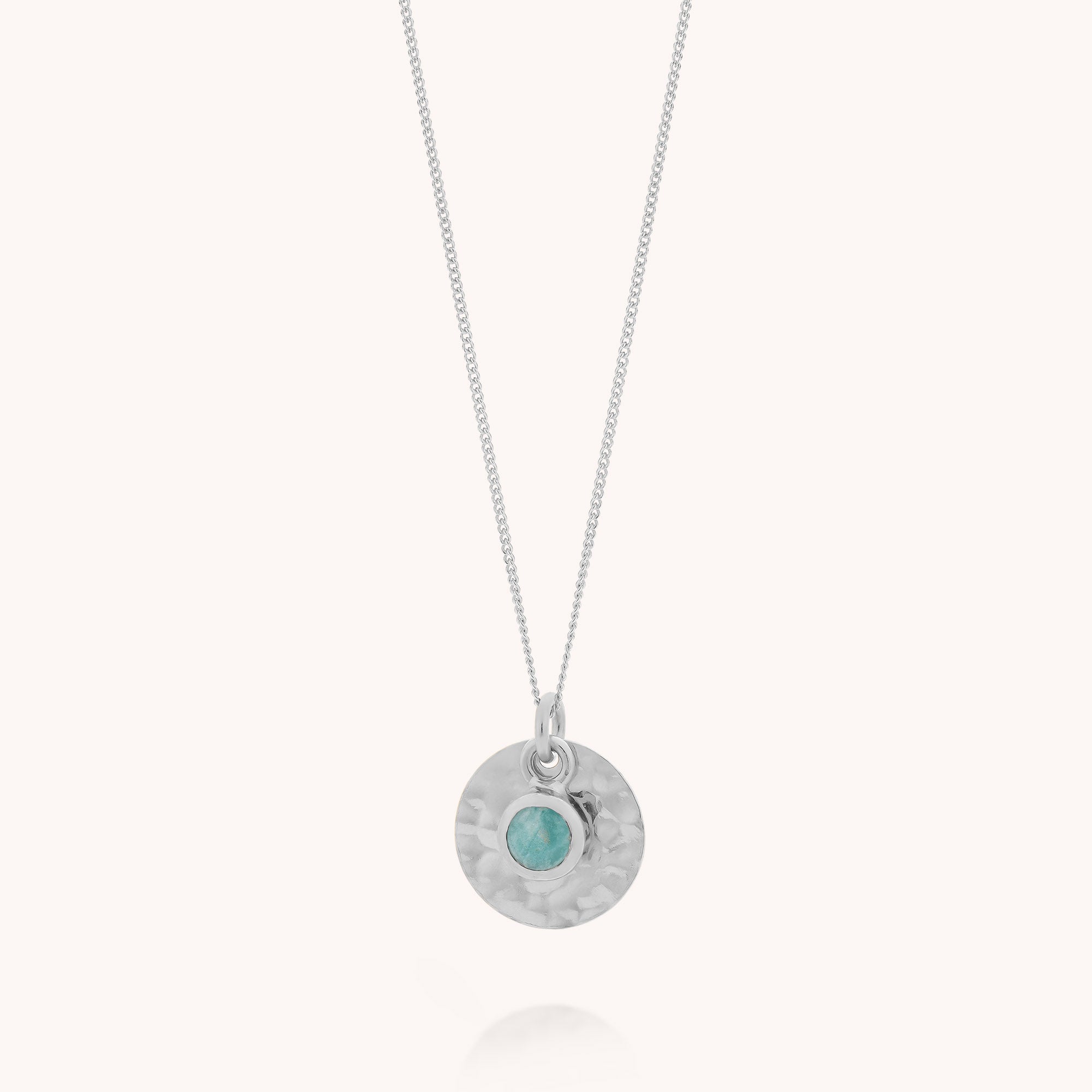 The Amazonite Luna Disc Necklace is a silver necklace with a fine chain, showcasing a round, hammered disc pendant inspired by jewelry design and accented with a small, turquoise-colored amazonite gemstone at the center.