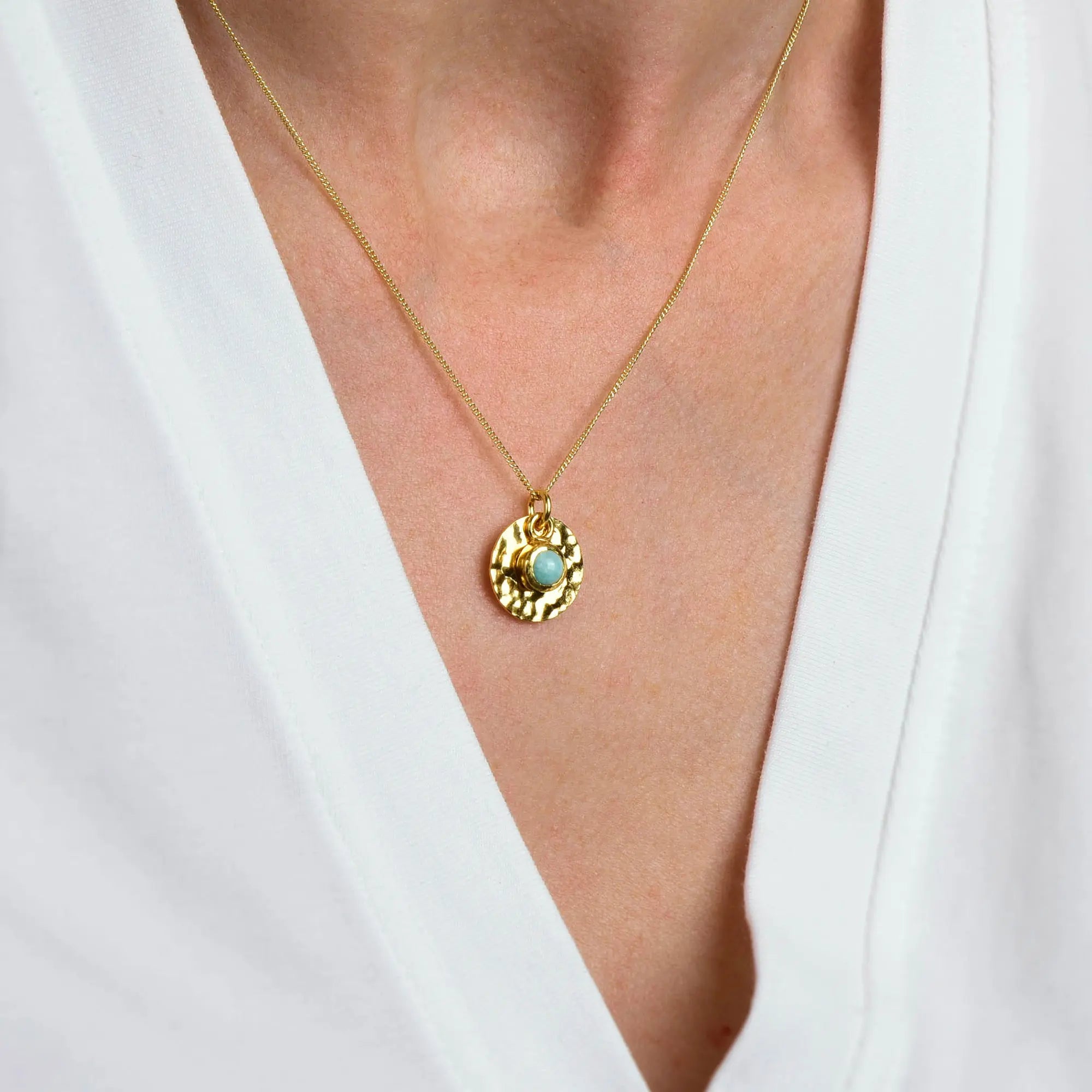 A close-up of a person wearing the Amazonite Luna Disc Necklace, featuring a round, hammered gold disc pendant with an amazonite stone, shown against a white V-neck shirt.