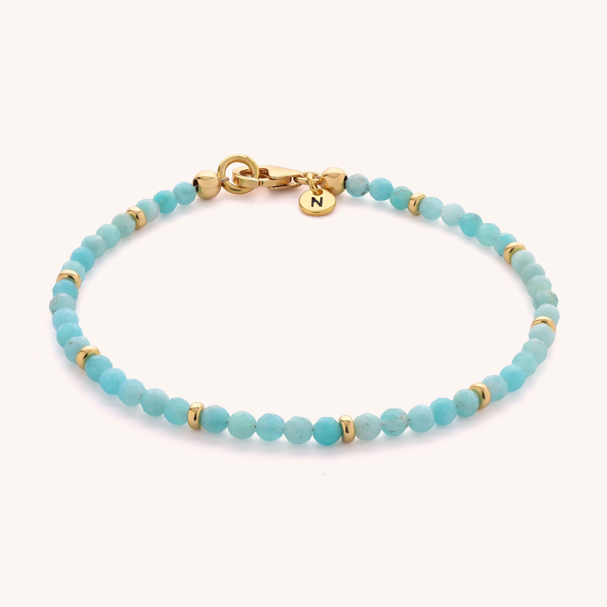 Introducing the Amazonite Orbit Bracelet: a delicate sterling silver bracelet adorned with petite turquoise and amazonite beads, accented with gold details. It features a gold-plated eco sterling silver clasp and is finished with a round gold charm engraved with the letter N.