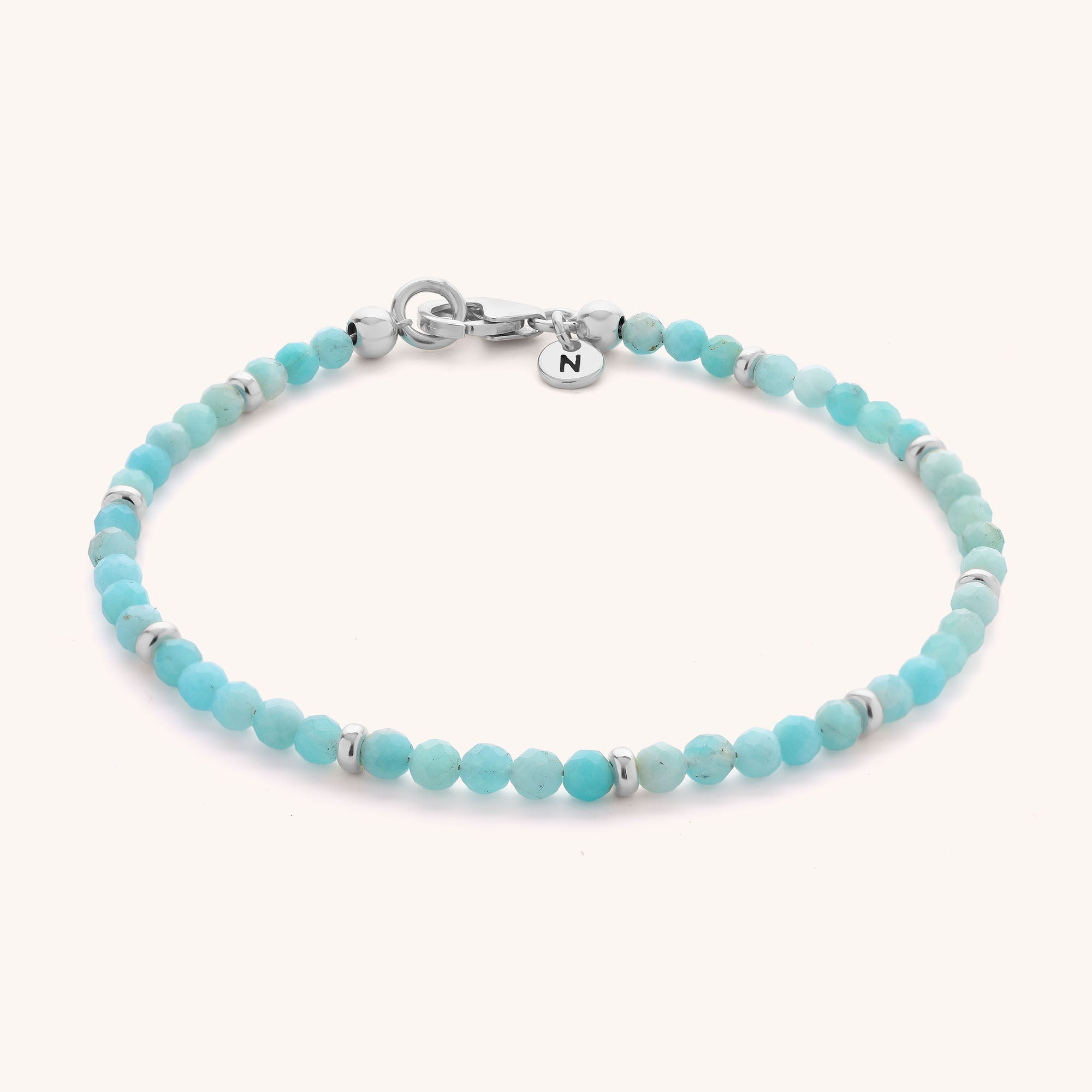 The Amazonite Orbit Bracelet is a delicate sterling silver bracelet featuring small round turquoise and amazonite beads with silver spacers, finished with a silver clasp and a round charm engraved with the letter "N.