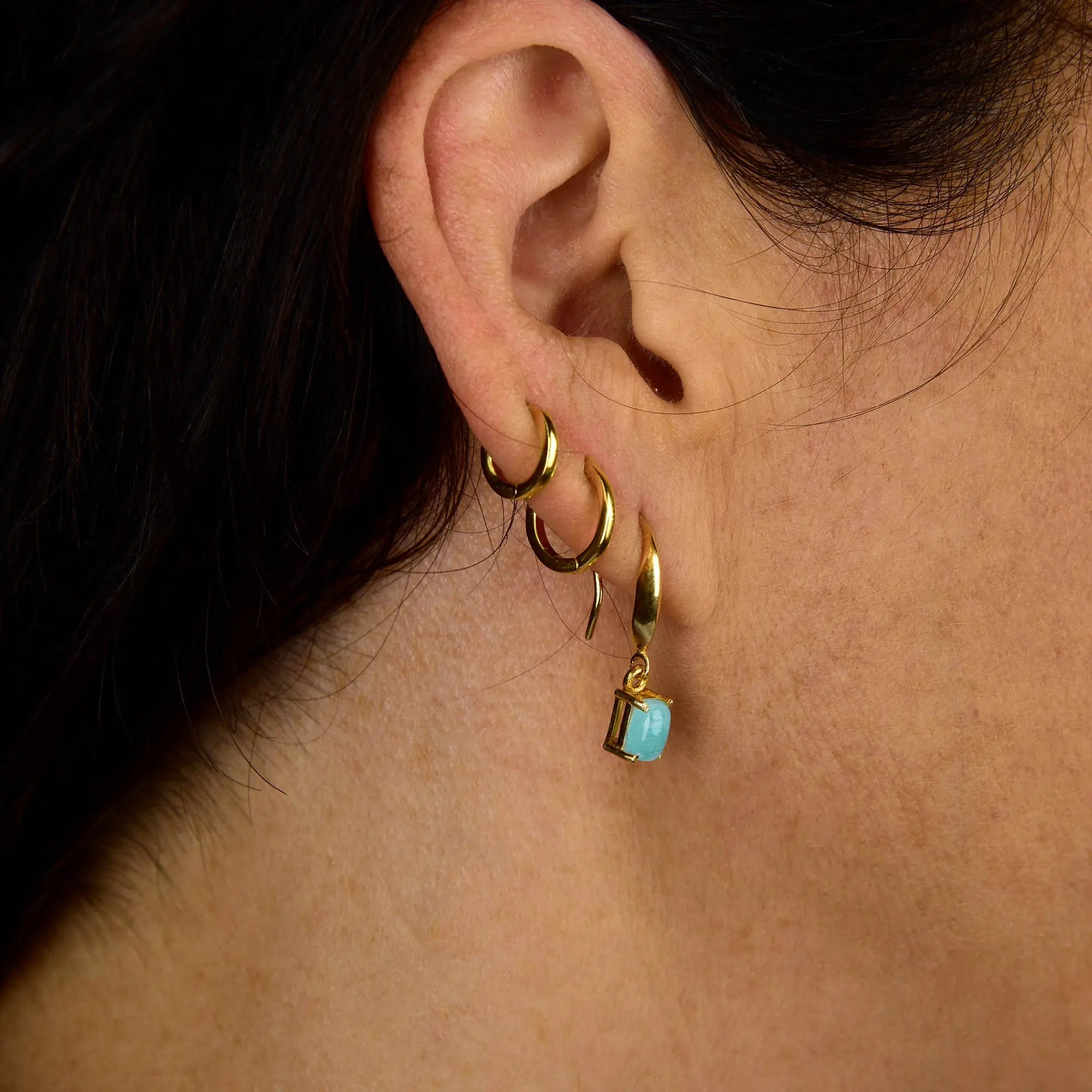 Close-up of a person’s ear adorned with three small gold hoop earrings and one Amazonite Octavia Drop Earring featuring a rectangular blue gemstone pendant in an octagonal gold setting. The person’s dark hair is pulled back, highlighting the ear and jewelry.