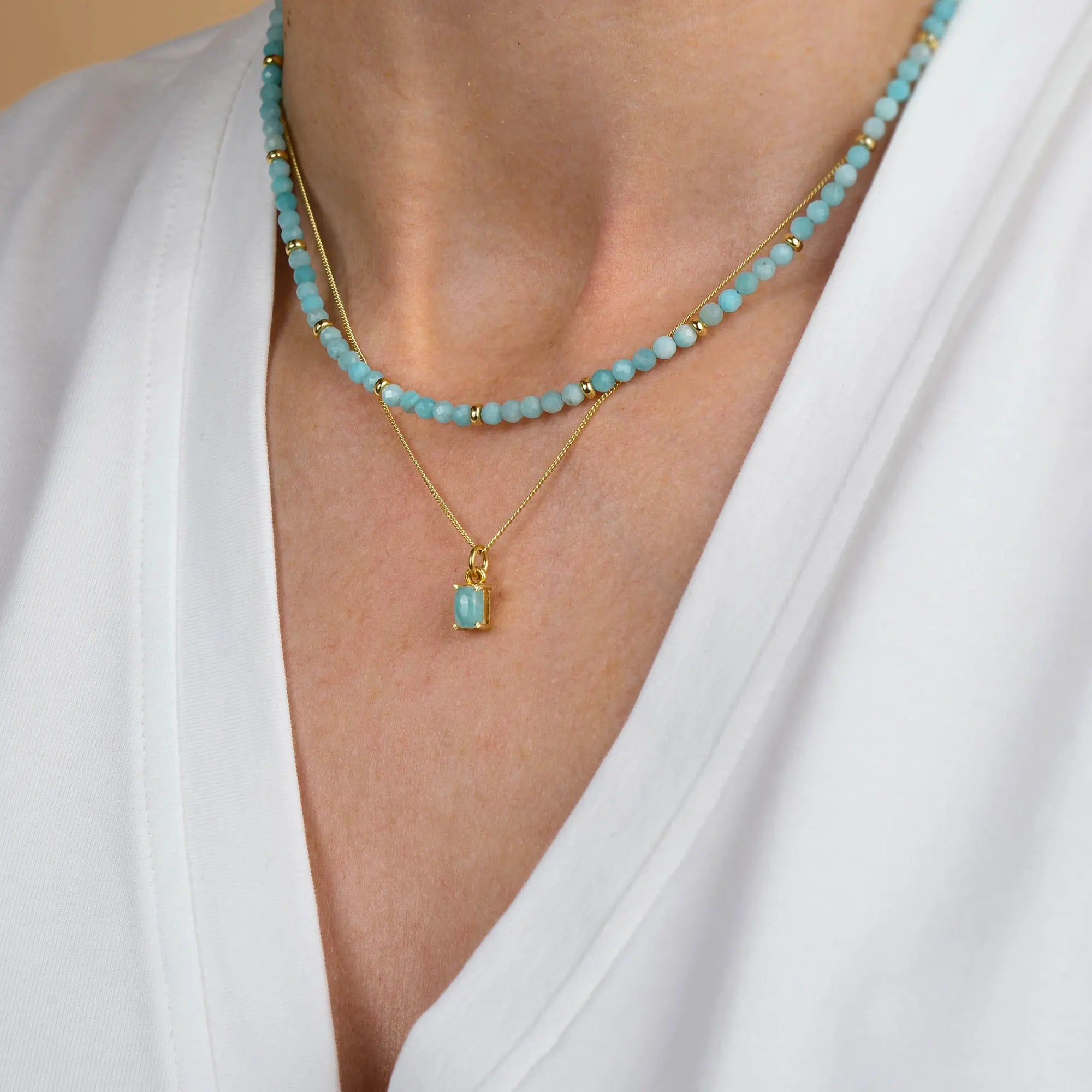 A person wearing a white V-neck shirt, a turquoise beaded necklace crafted with natural gemstones, and the Amazonite Orbit Necklace—a gold chain featuring a rectangular amazonite pendant—beautiful examples of handcrafted jewellery.