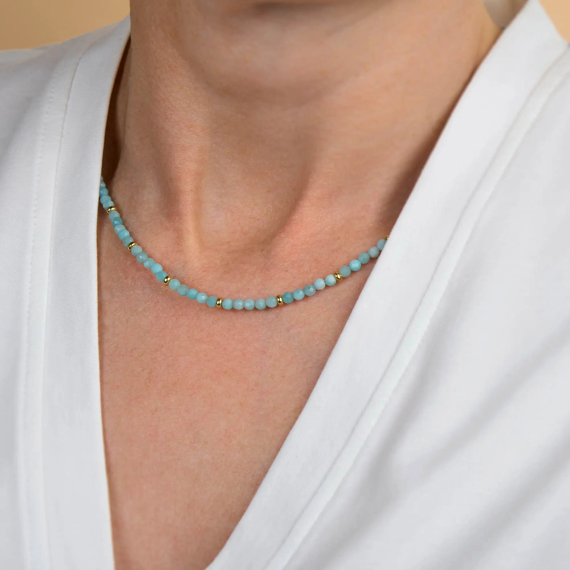A person wearing a white v-neck top and the Amazonite Orbit Necklace—a delicate handcrafted jewellery piece featuring light blue natural gemstones and small gold beads—around their neck.