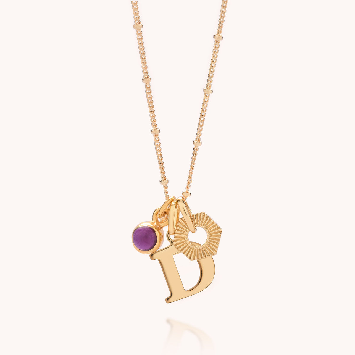 Presenting the HEX Personalised Birthstone Necklace – Gold Plated: a stunning necklace crafted from gold-plated sterling silver, featuring a delicate chain with small bead accents. Showcasing three unique charms—a round purple birthstone gem, a sunburst design, and a gold letter D—this beautiful piece makes an ideal personalised gift. Photographed against a plain, light-colored background.