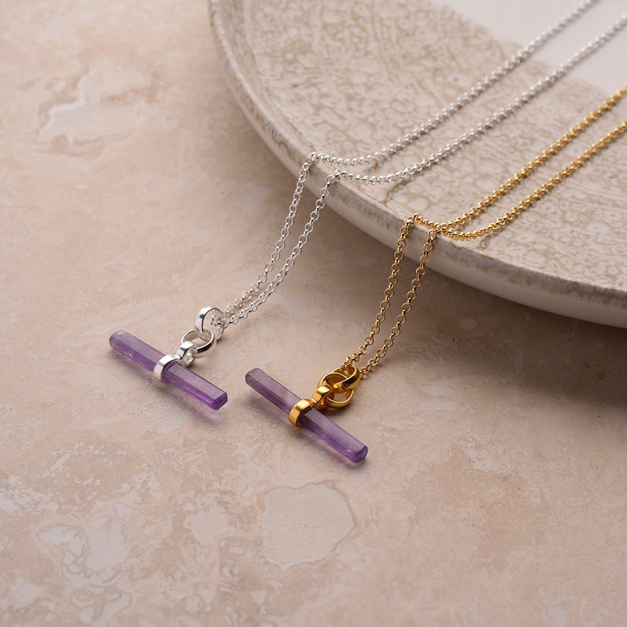 Two Gemstone T Bar Necklaces—one with a sterling silver chain, the other with a gold chain—feature horizontal purple gemstone pendants and are displayed on a beige textured surface next to a round ceramic plate.