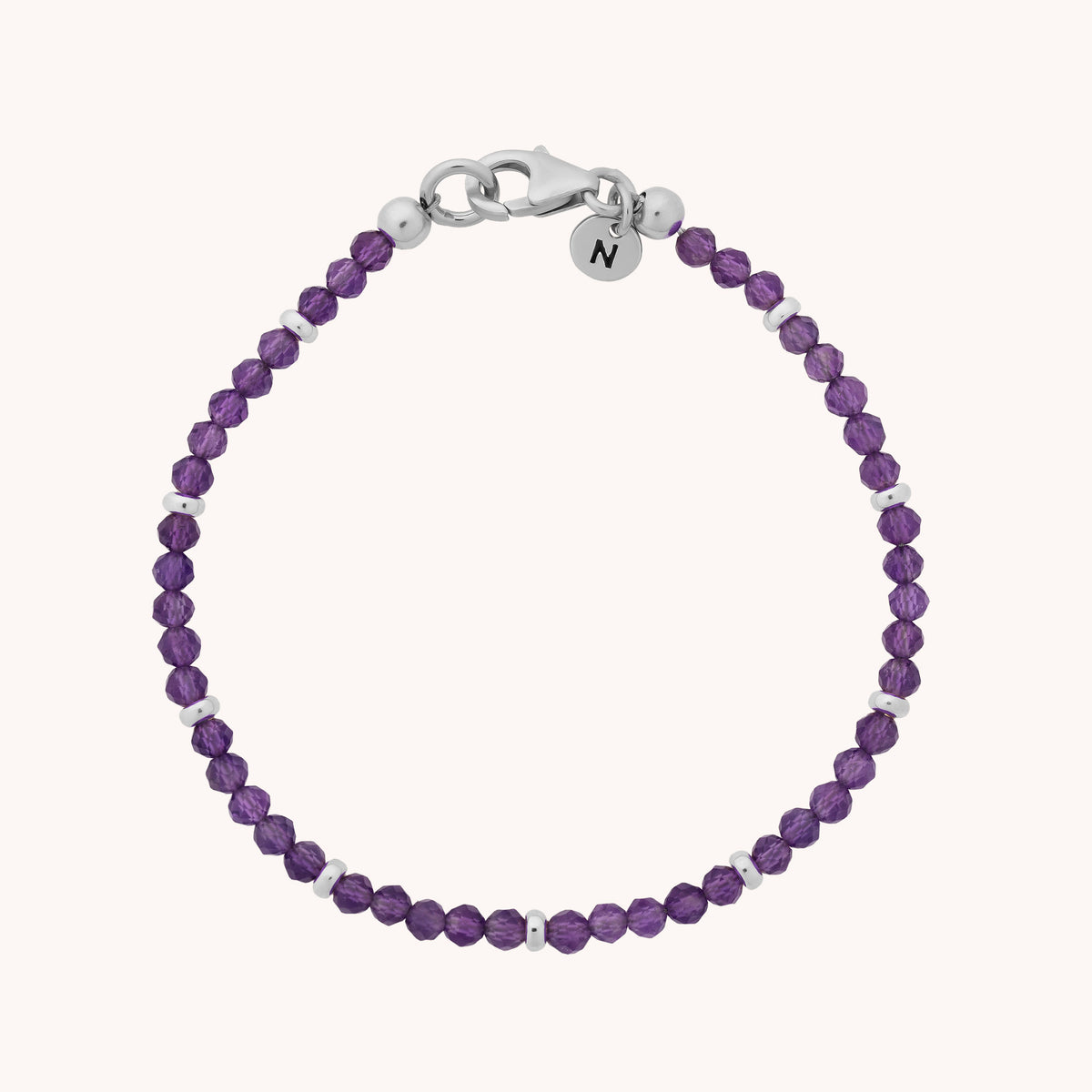 The Amethyst Orbit Bracelet - February features small, faceted purple amethyst beads with sterling silver accents, a silver clasp, and a round tag engraved with the letter N, beautifully displayed against a light background.