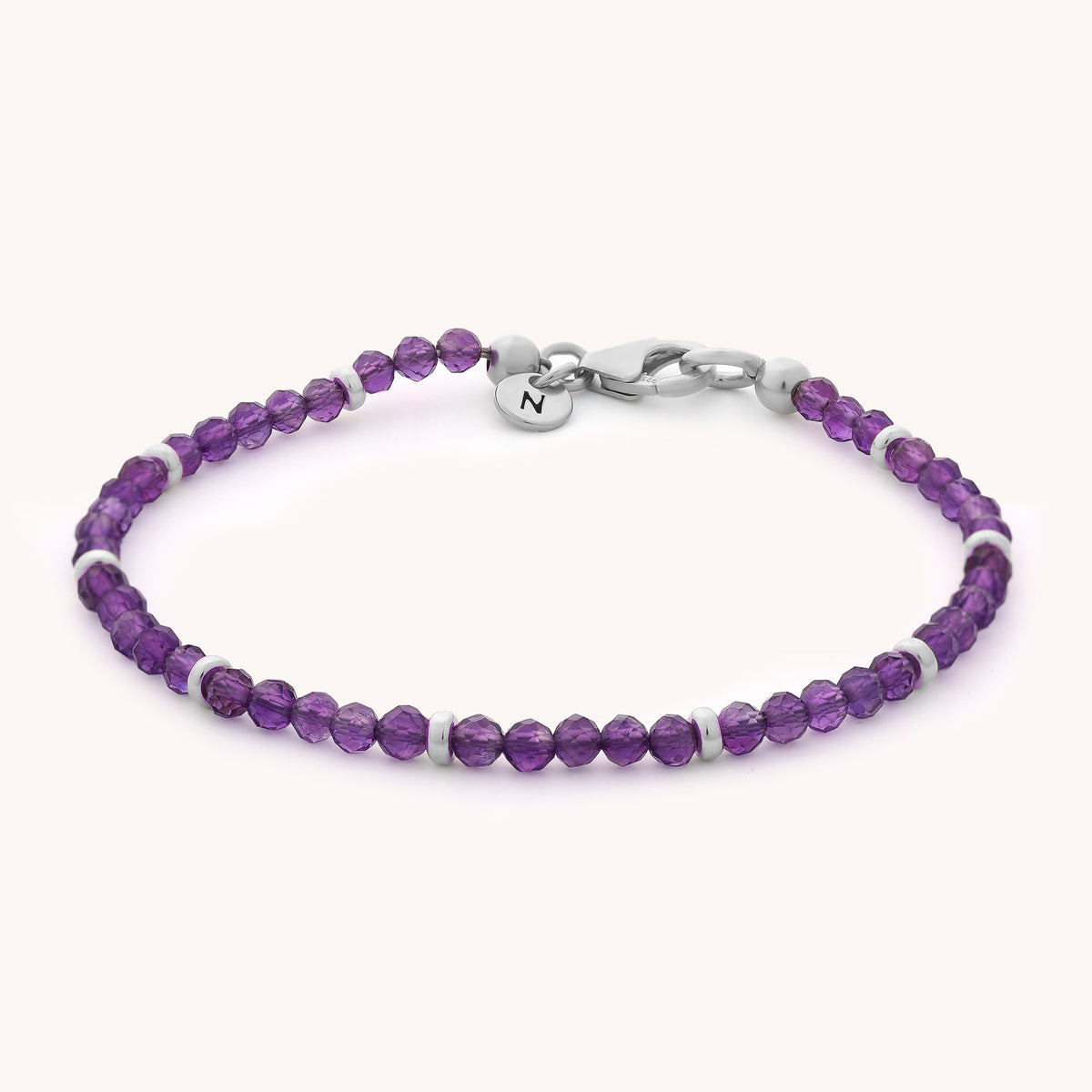 The Amethyst Orbit Bracelet - February showcases delicate, faceted purple amethyst beads accented with sterling silver spacers and a metal clasp, finished with a round charm engraved with the letter N—all beautifully inspired by the February birthstone.