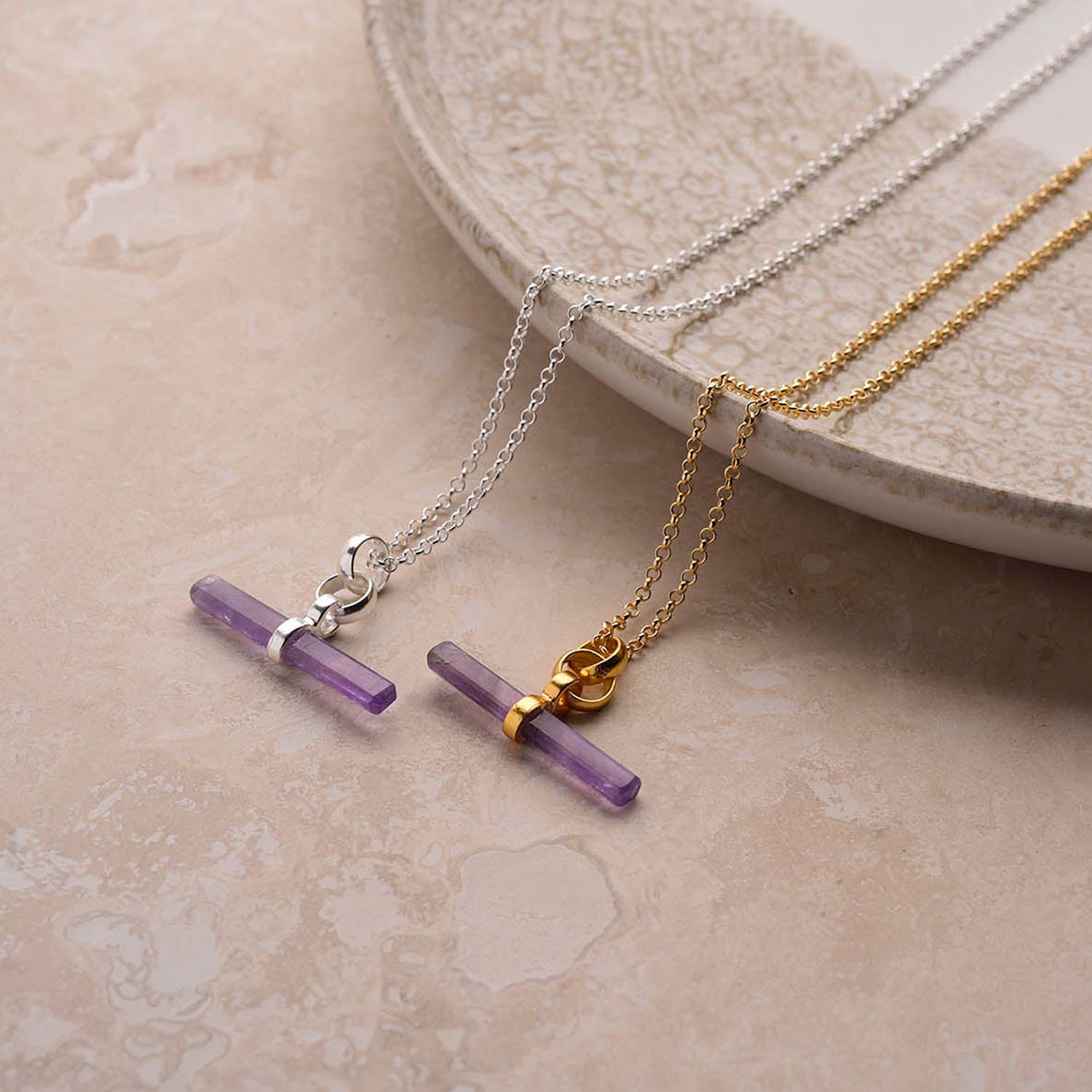 Two Gemstone T Bar Necklace - Amethyst pieces—one on a sterling silver chain, one on gold—are displayed on beige marble next to a ceramic plate, each showcasing a slim purple amethyst pendant.