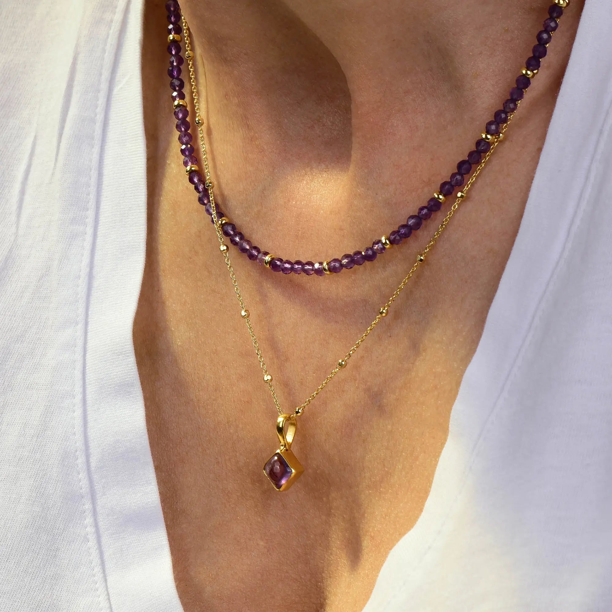 A person wearing a white V-neck top, a beaded purple necklace, and the Amethyst February Birthstone Orbit Necklace—a gold handcrafted piece adorned with a vibrant purple gemstone pendant that beautifully highlights the February birthstone.