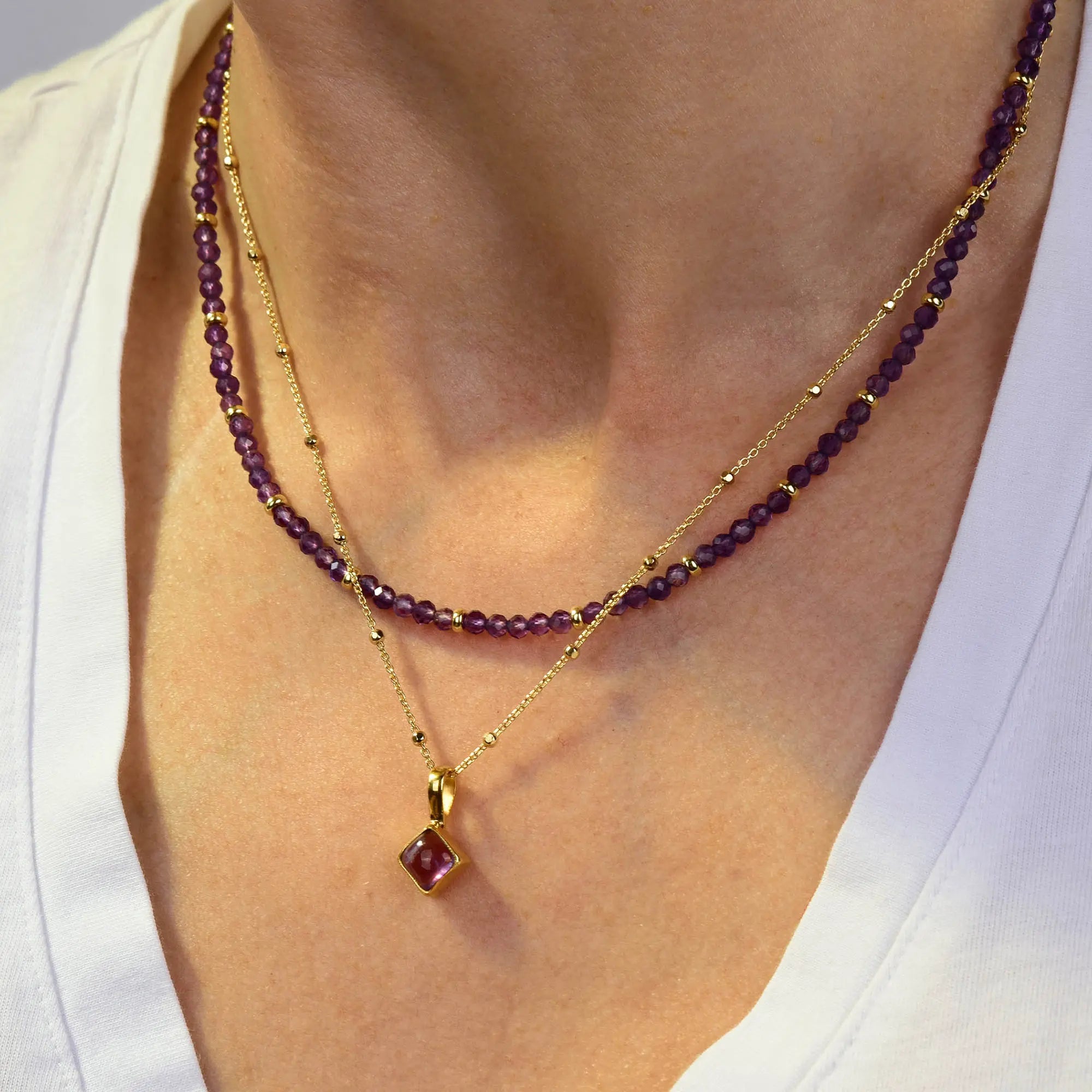 A person wearing two gold necklaces: one with small purple beads and the other, the Amethyst February Birthstone Orbit Necklace, featuring a handcrafted gold pendant with an amethyst stone, over a white V-neck shirt.
