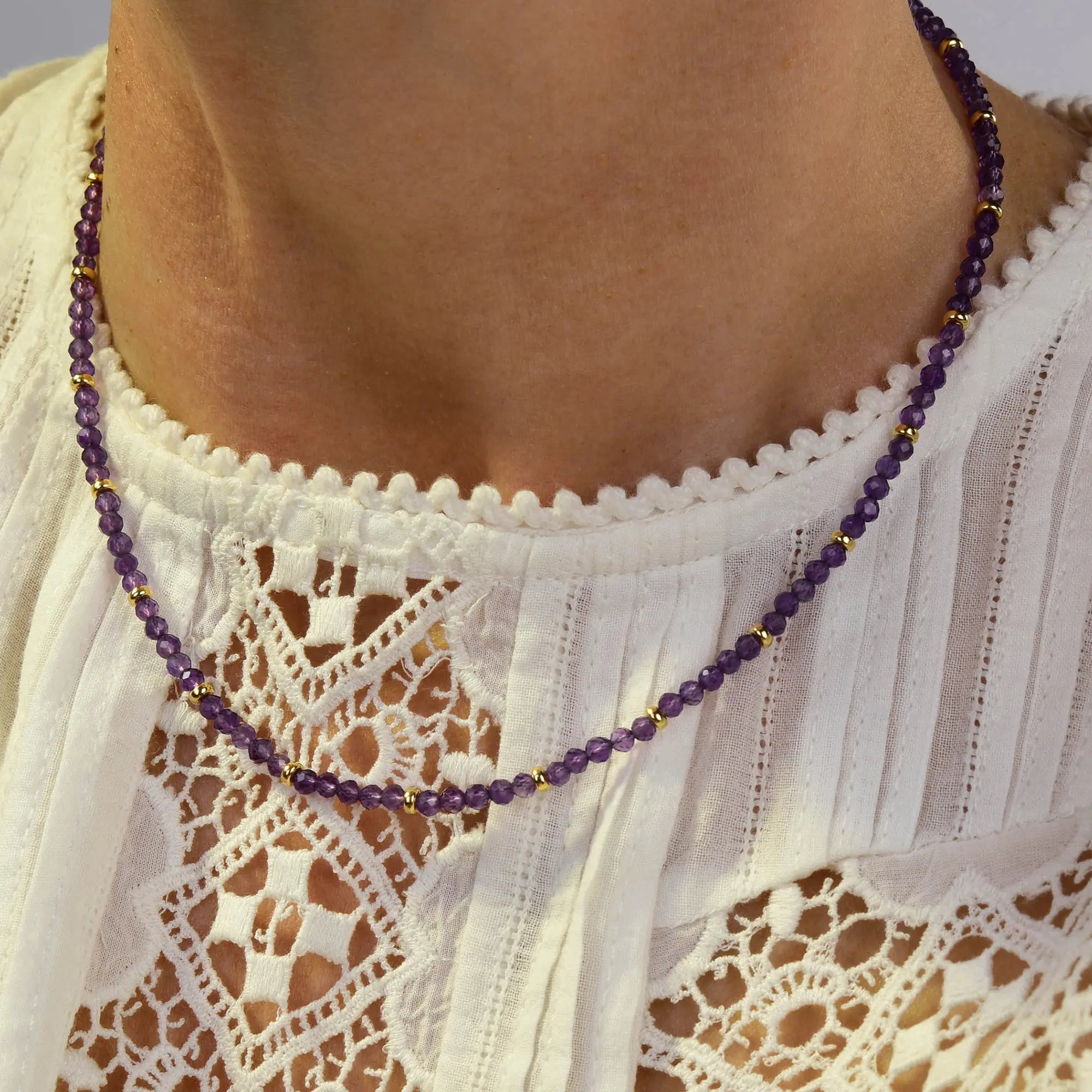 A close-up of a person wearing the Amethyst February Birthstone Orbit Necklace, featuring delicate purple amethyst beads and gold accents, styled over a white lace top with intricate patterns and scalloped edges. This handcrafted necklace beautifully highlights the timeless elegance of February’s birthstone.