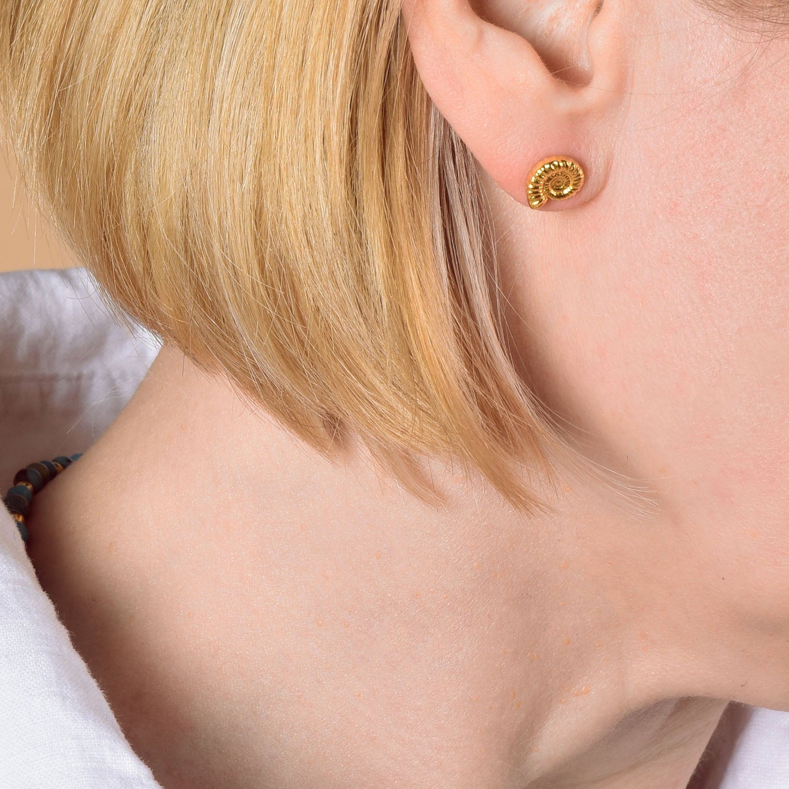 A woman with short blonde hair wears a white shirt, gold stud earring, and the Turquoise Jewel Bead Birthstone Necklace - December. Only the lower half of her face and neck appear against a beige background.