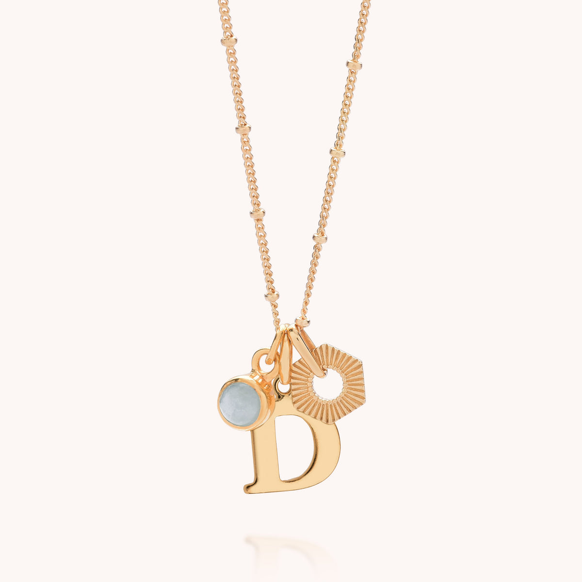 The HEX Personalised Birthstone Necklace – Gold Plated features a delicate gold-plated sterling silver chain adorned with three unique charms: a round light blue birthstone, a hexagonal sunburst design, and a gold letter D pendant. Set against a light background, this striking necklace makes a thoughtful personalised gift.
