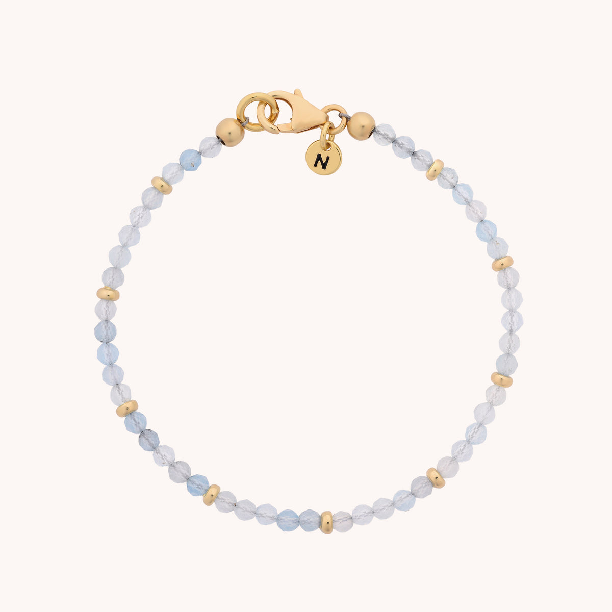 The Aquamarine Orbit Bracelet – March features light blue aquamarine beads reminiscent of the March birthstone, accented with gold spacers, a gold clasp, and a round gold charm engraved with the letter N. This piece combines gemstone jewelry elegance with personalized detail.