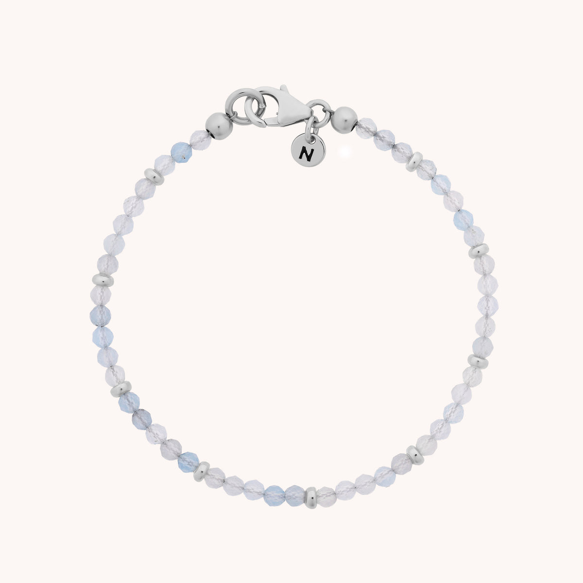 The Aquamarine Orbit Bracelet - March showcases translucent light blue aquamarine beads reminiscent of the March birthstone, accented by small silver spacers, a secure lobster clasp, and a round charm engraved with the letter N. Displayed on a light background, this bracelet makes for an elegant and charming piece of gemstone jewelry.
