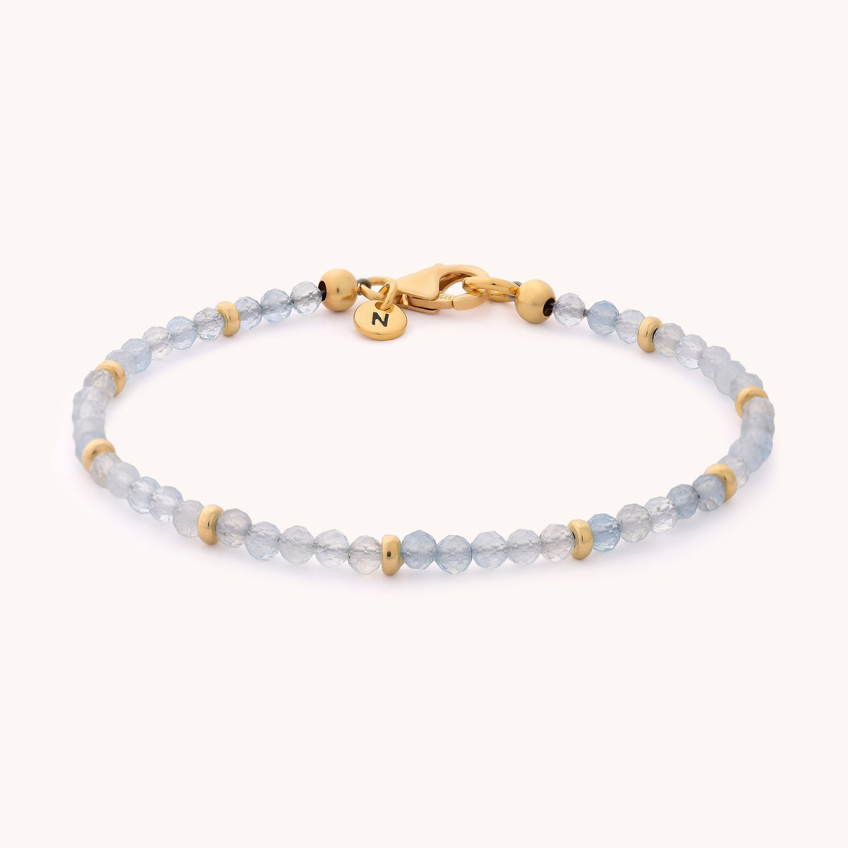 Introducing the Aquamarine Orbit Bracelet - March: a delicate piece adorned with small light blue aquamarine beads, spaced by tiny gold-colored accents, and finished with a gold clasp featuring a round charm engraved with the letter N. This gemstone bracelet makes an ideal gift for anyone celebrating a March birthstone.