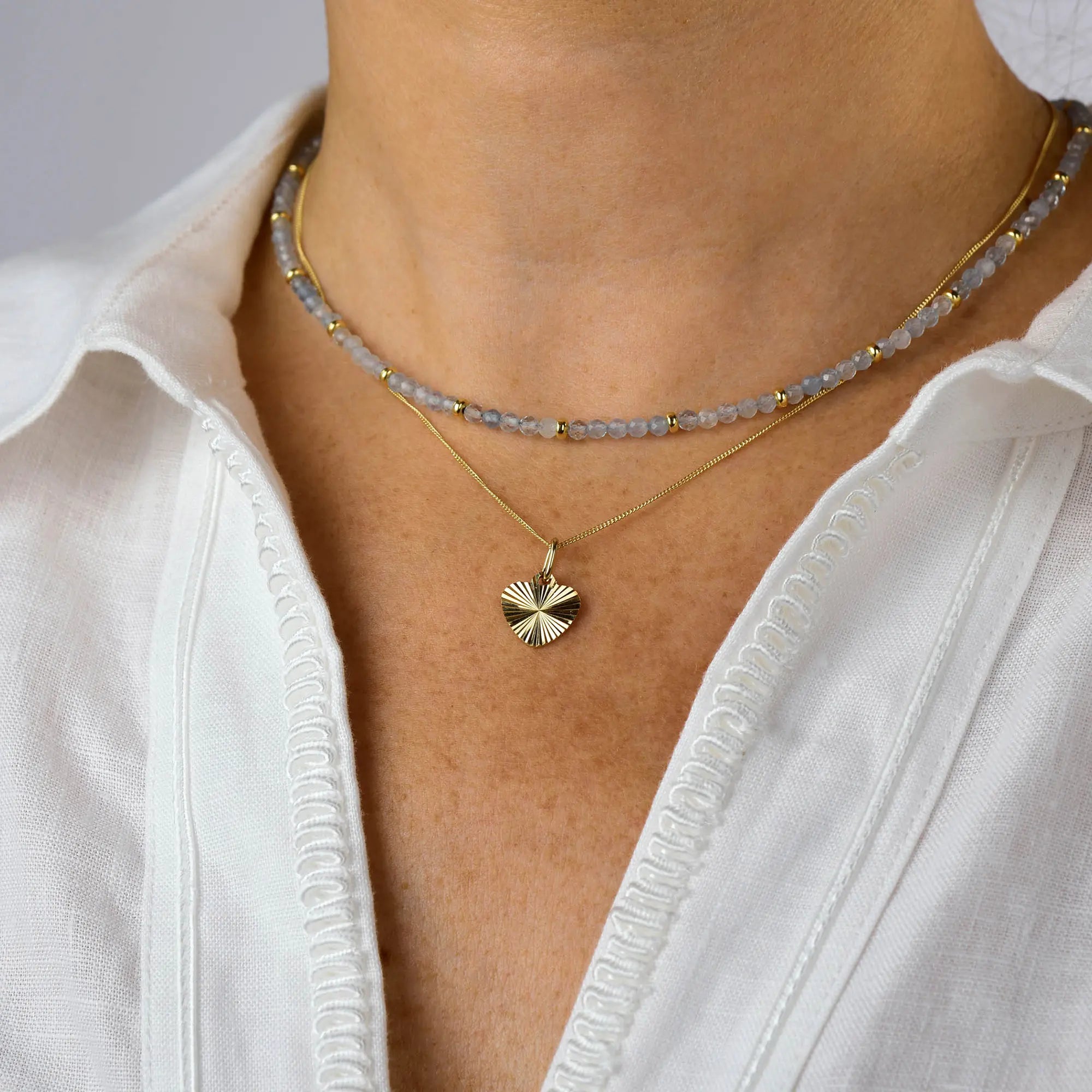 A person wearing a white embroidered shirt, a beaded choker necklace, and the Celeste Heart Necklace—a gold plated pendant shaped like a heart.