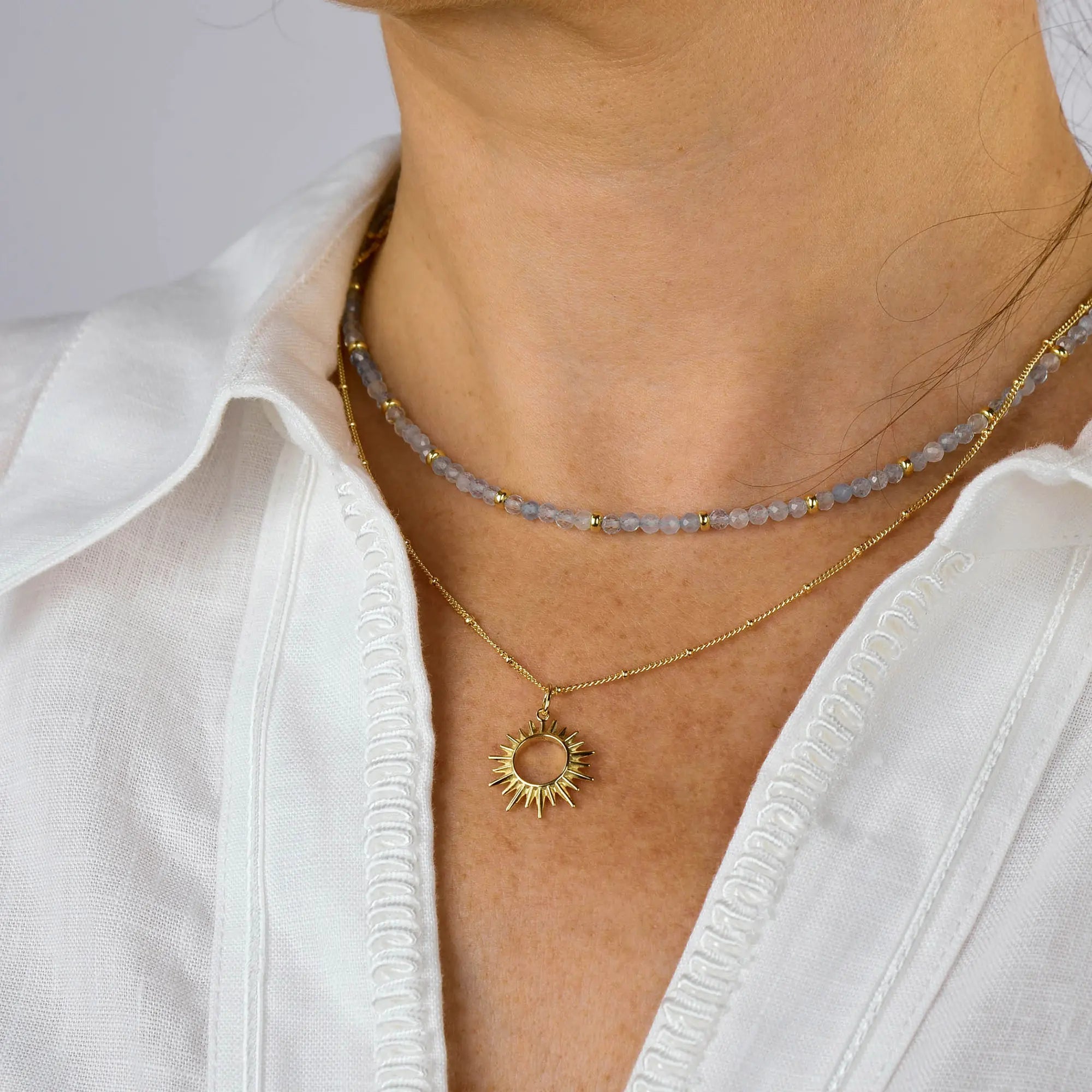 A person wearing a white button-up shirt and two necklaces: one is a beaded choker, and the other is the Sol Necklace—a gold-plated piece featuring a sun-shaped pendant.