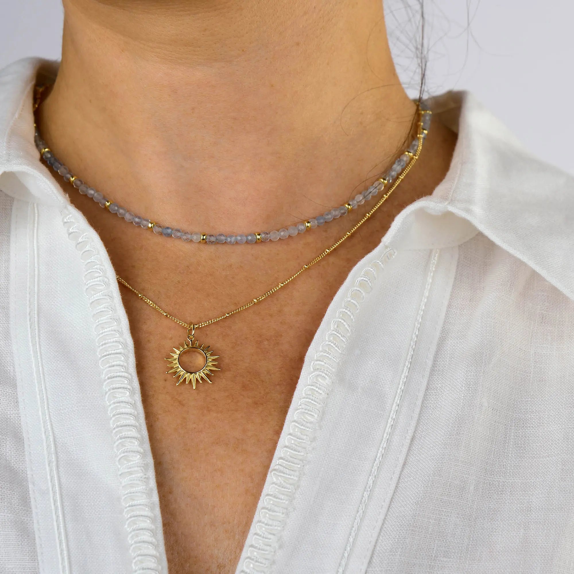A person wearing a white shirt with two necklaces: the Sol Necklace, a gold-plated necklace featuring a sun pendant, and a beaded choker with gold and gray beads.