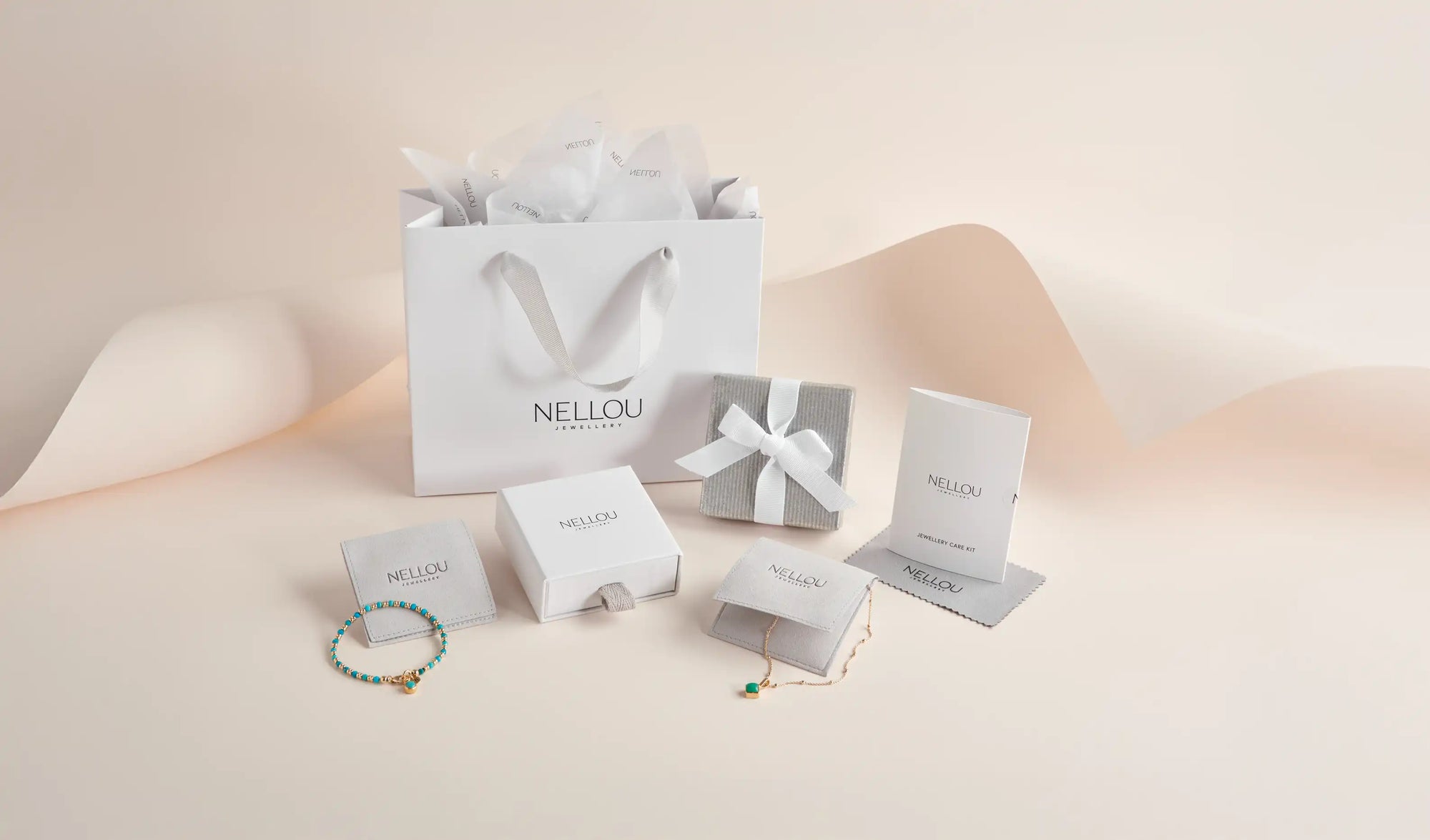 A collection of white and gray NELLOU-branded jewelry packaging, including a gift bag, boxes, pouches, and cards, displayed alongside a turquoise beaded bracelet and a gold necklace with a green heart charm.