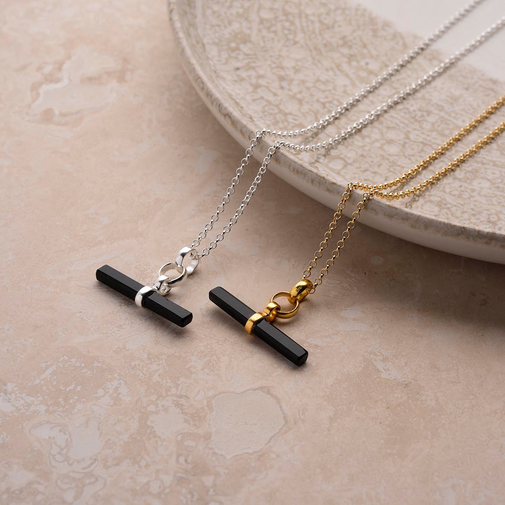 Two Gemstone T Bar Necklaces with black gemstone bars lie parallel on a beige marble surface—one features a silver chain and clasp, the other gold—set near the edge of a ceramic plate.