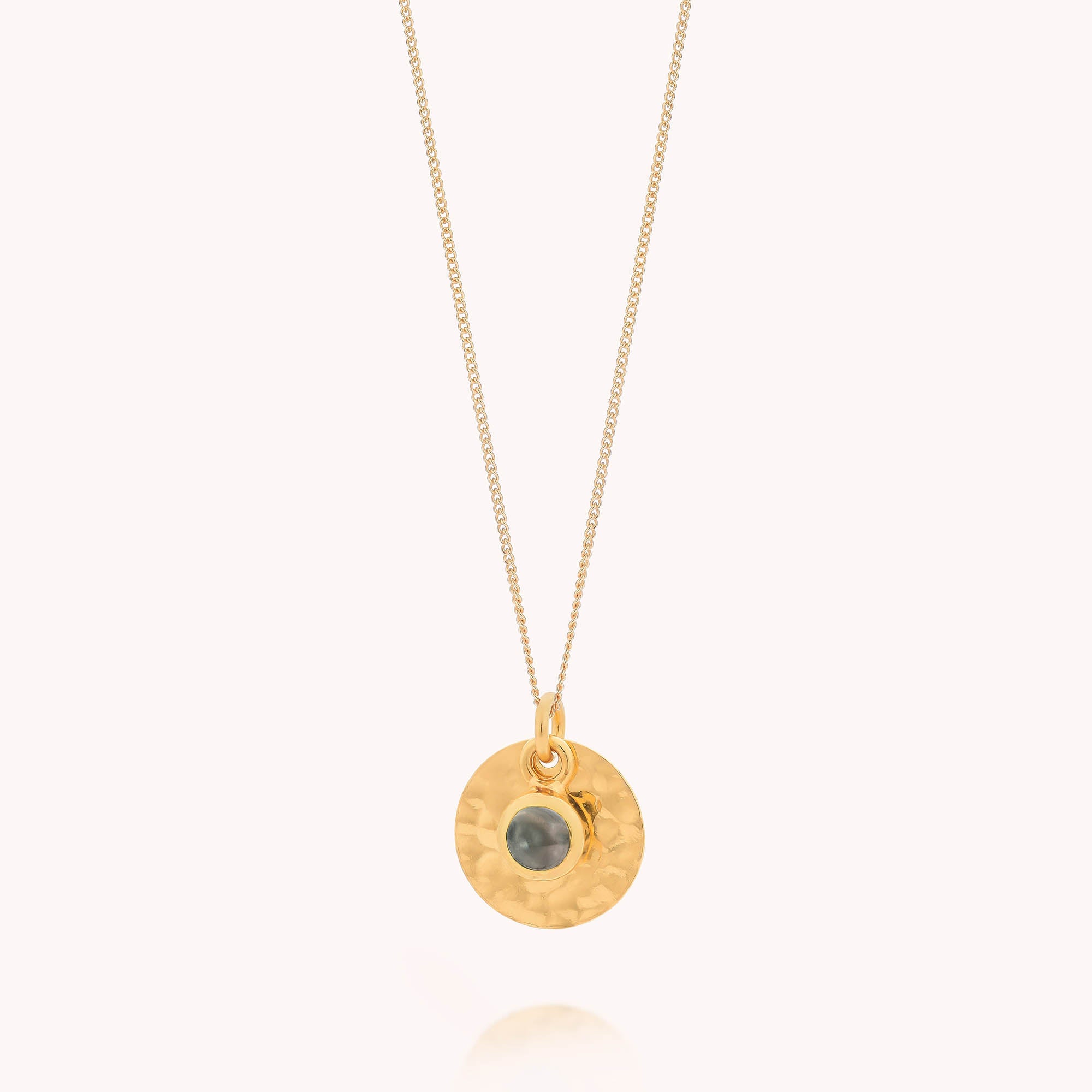 The Blue Topaz Luna Disc Necklace features a thin gold chain with a round, hammered disc pendant. A small blue topaz stone, representing the December birthstone, is set at the center and displayed on a simple light background.
