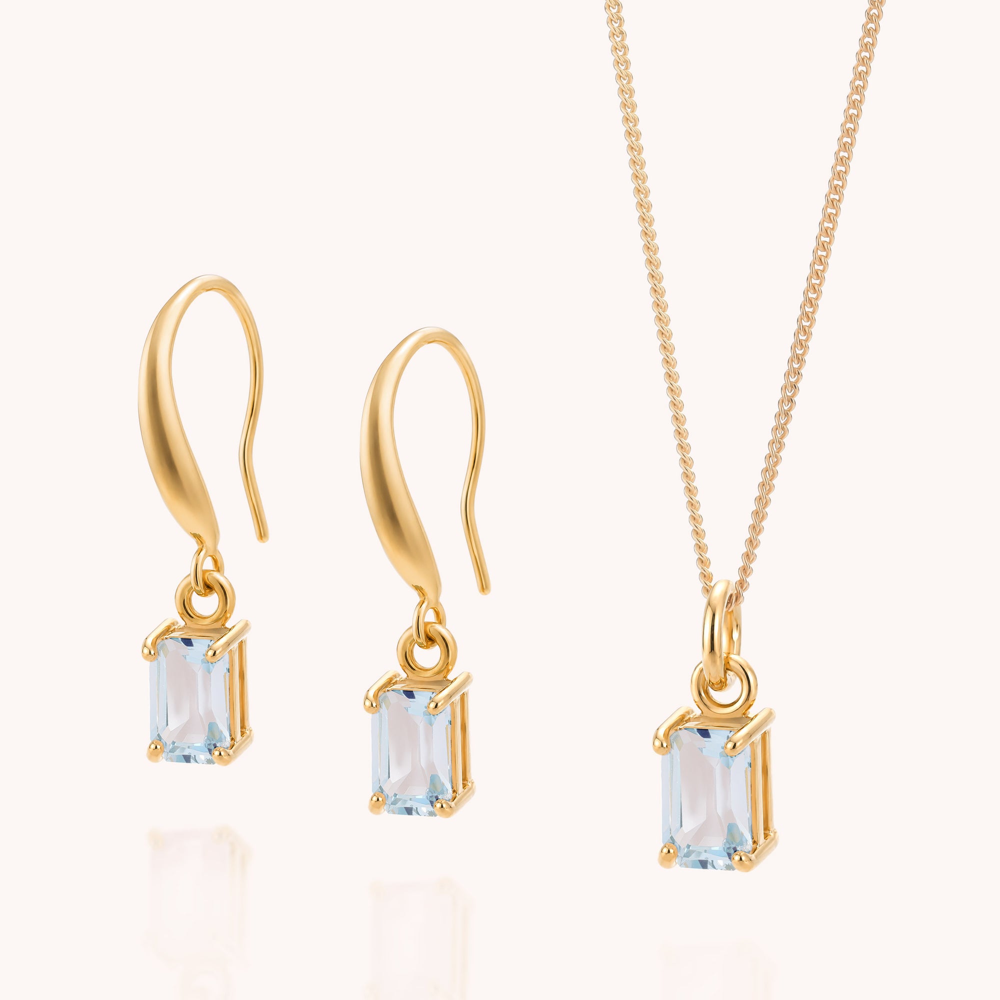 The Blue Topaz Octavia Necklace &amp; Earrings Set features a matching gold jewelry ensemble with two drop earrings and a necklace, each adorned with a rectangular light blue topaz gemstone—evoking the elegance of the December birthstone—securely set in refined gold prongs and showcased against a light background.
