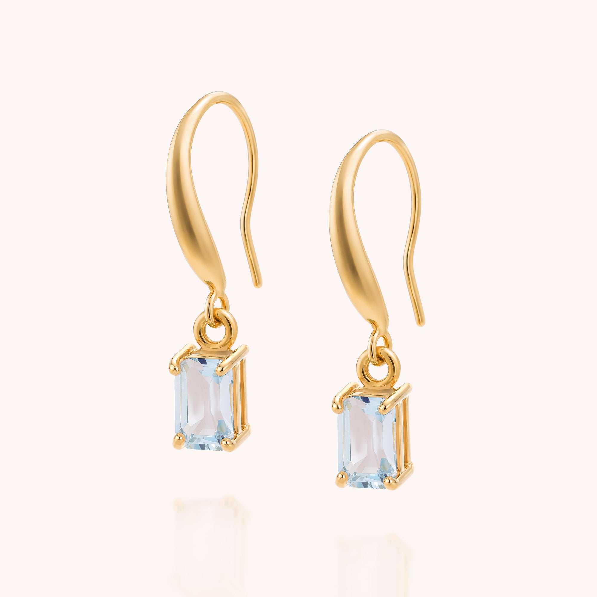 Gold drop earrings featuring rectangular, light blue gemstones in prong settings on a plain, light background—these striking Blue Topaz Octavia Drop Earrings make a stunning December birthstone choice.