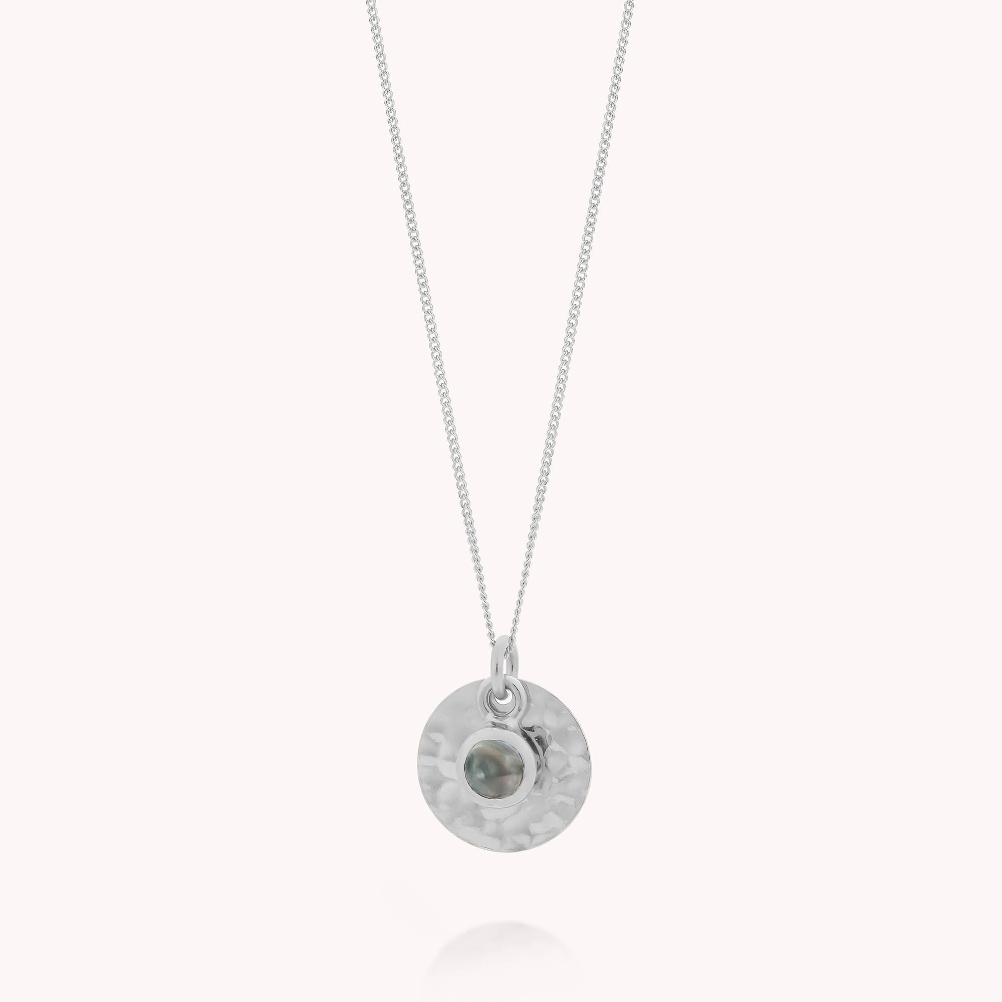 The Blue Topaz Luna Disc Necklace features a delicate sterling silver chain with a round, hammered pendant accented by a small blue topaz gemstone—an elegant accessory and perfect December birthstone gift.