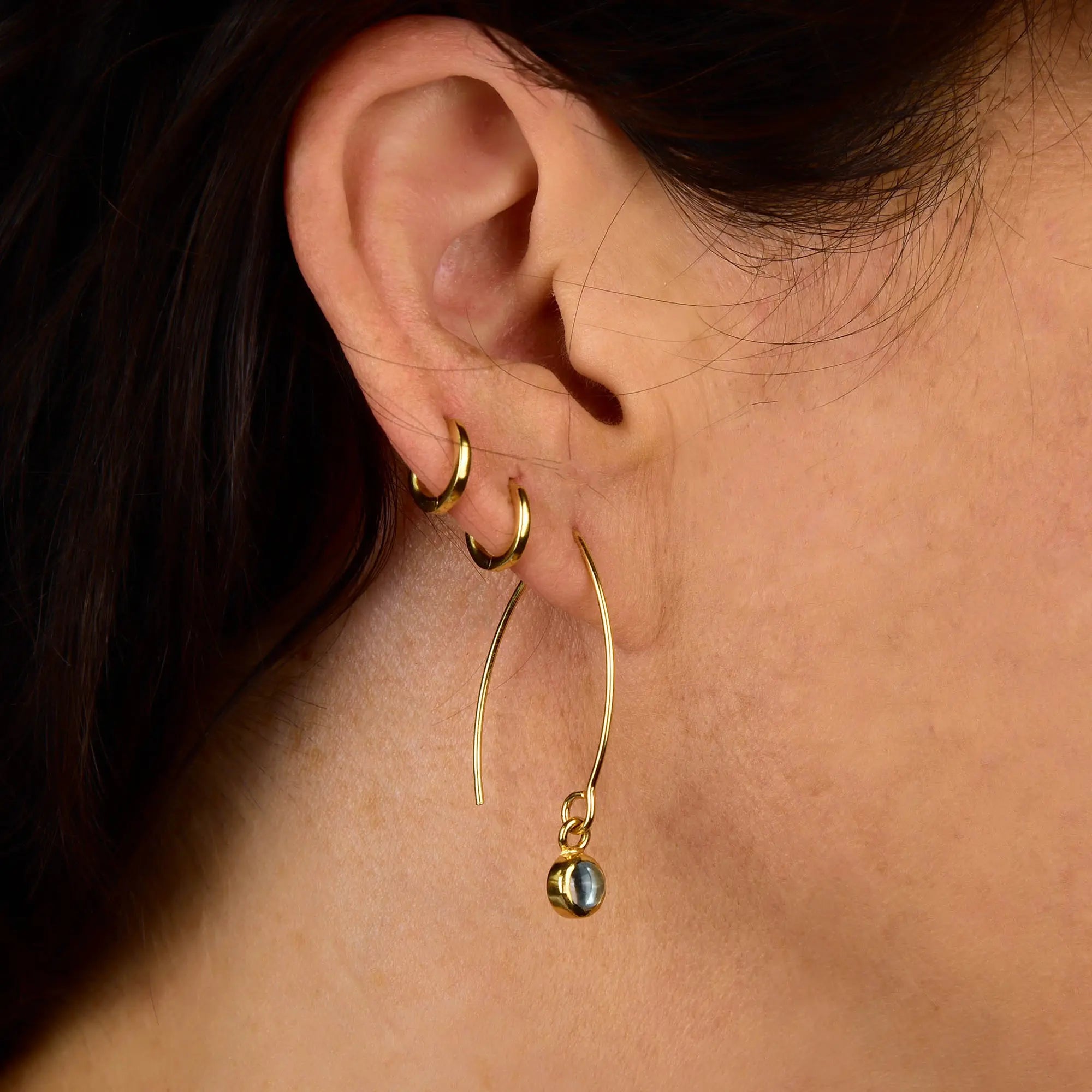 Close-up of a person’s ear adorned with three gold earrings: two small hoops on the upper lobe and one long hoop featuring a dangling sphere on the lower lobe. With dark hair tucked neatly behind the ear, this elegant arrangement highlights each piece—a beautiful way to showcase gemstone accents like the Blue Topaz Gem Dot Arc Earrings, making them perfect for December birthstone celebrations.