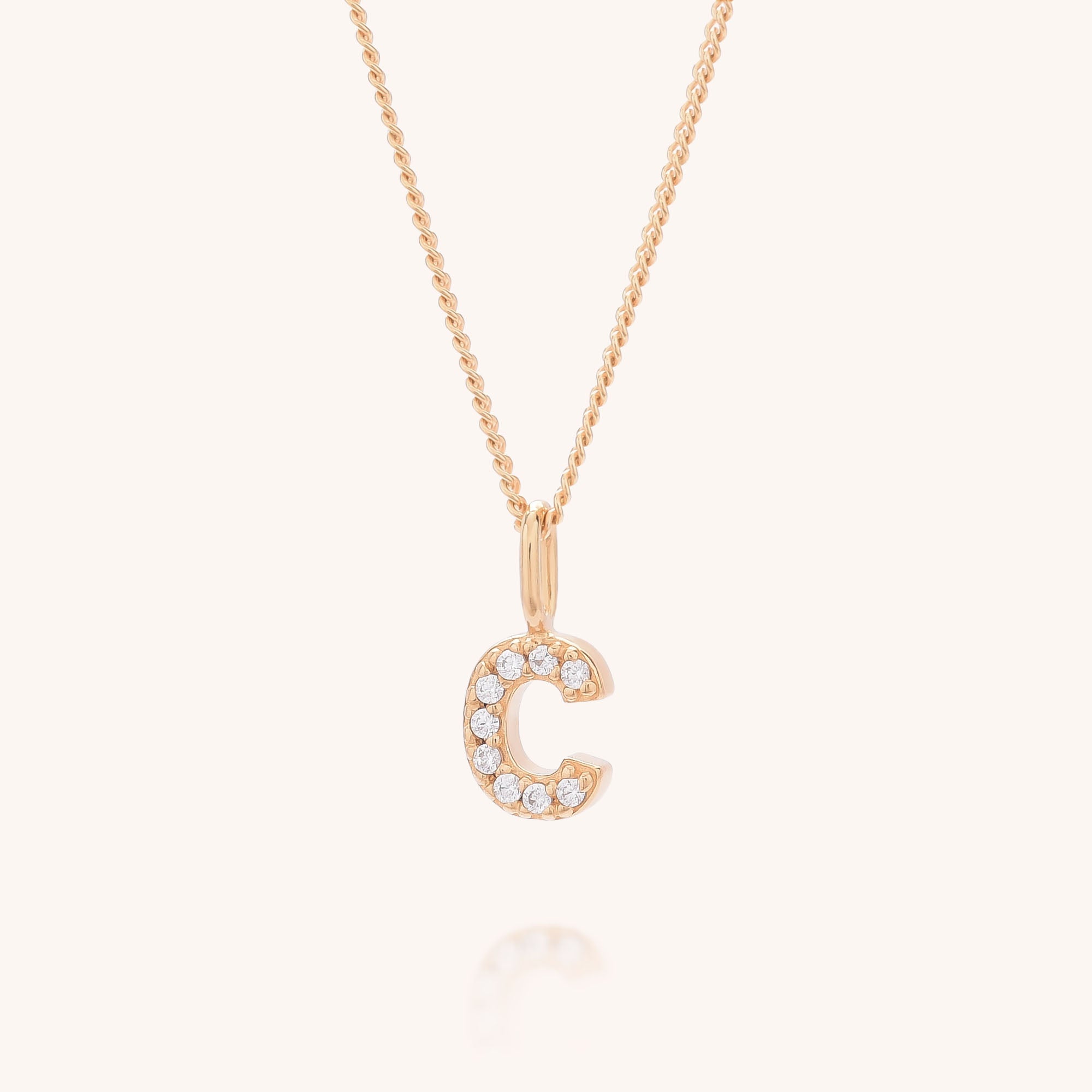 The Initial Sparkle Necklace is a gold chain with a letter C pendant, set with sparkling cubic zirconia stones, shown on a soft light beige background.