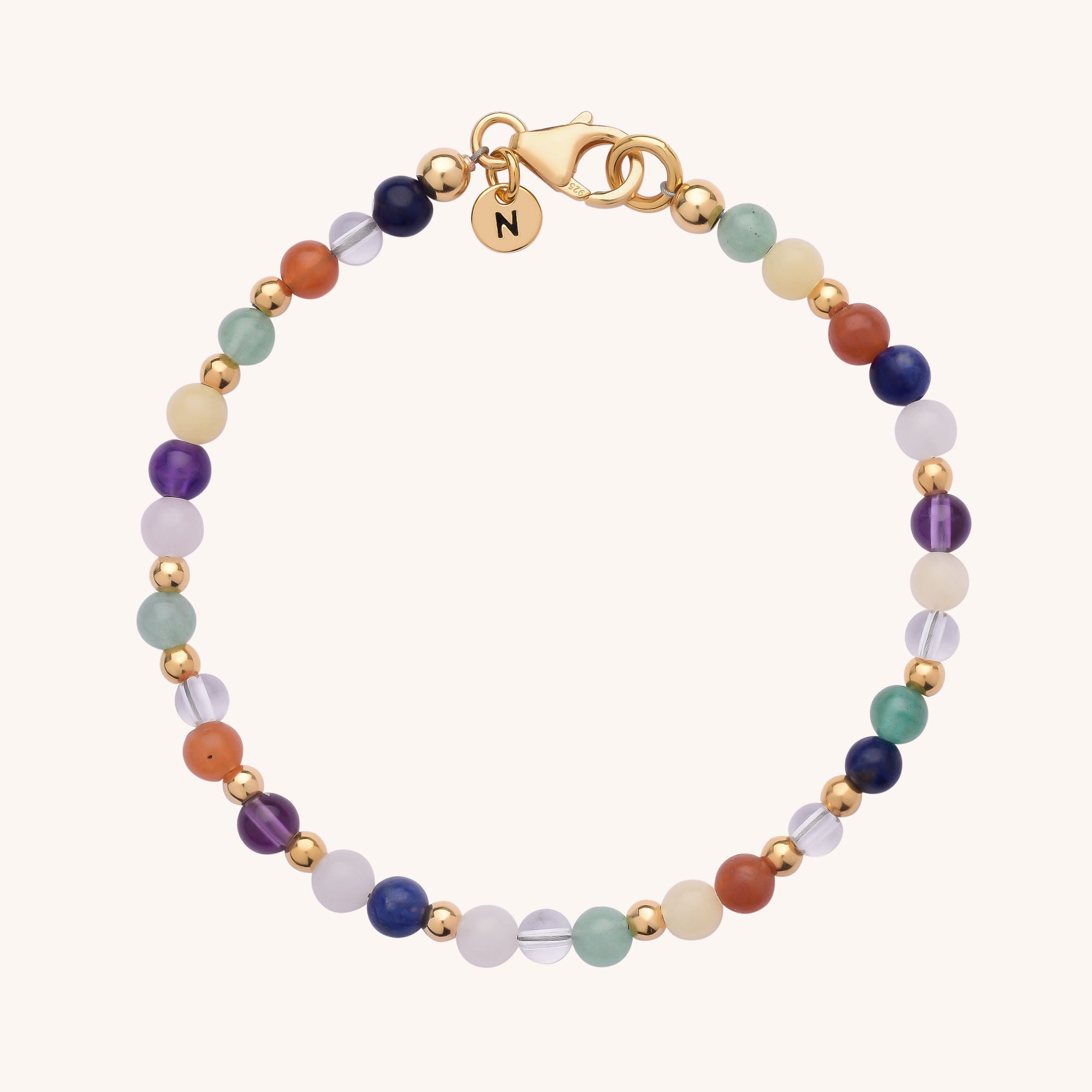 The Jewel Bead Chakra Bracelet features multicolored round natural gemstone beads in green, purple, blue, orange, and white with gold accents and a gold clasp adorned with a round charm engraved with the letter N.