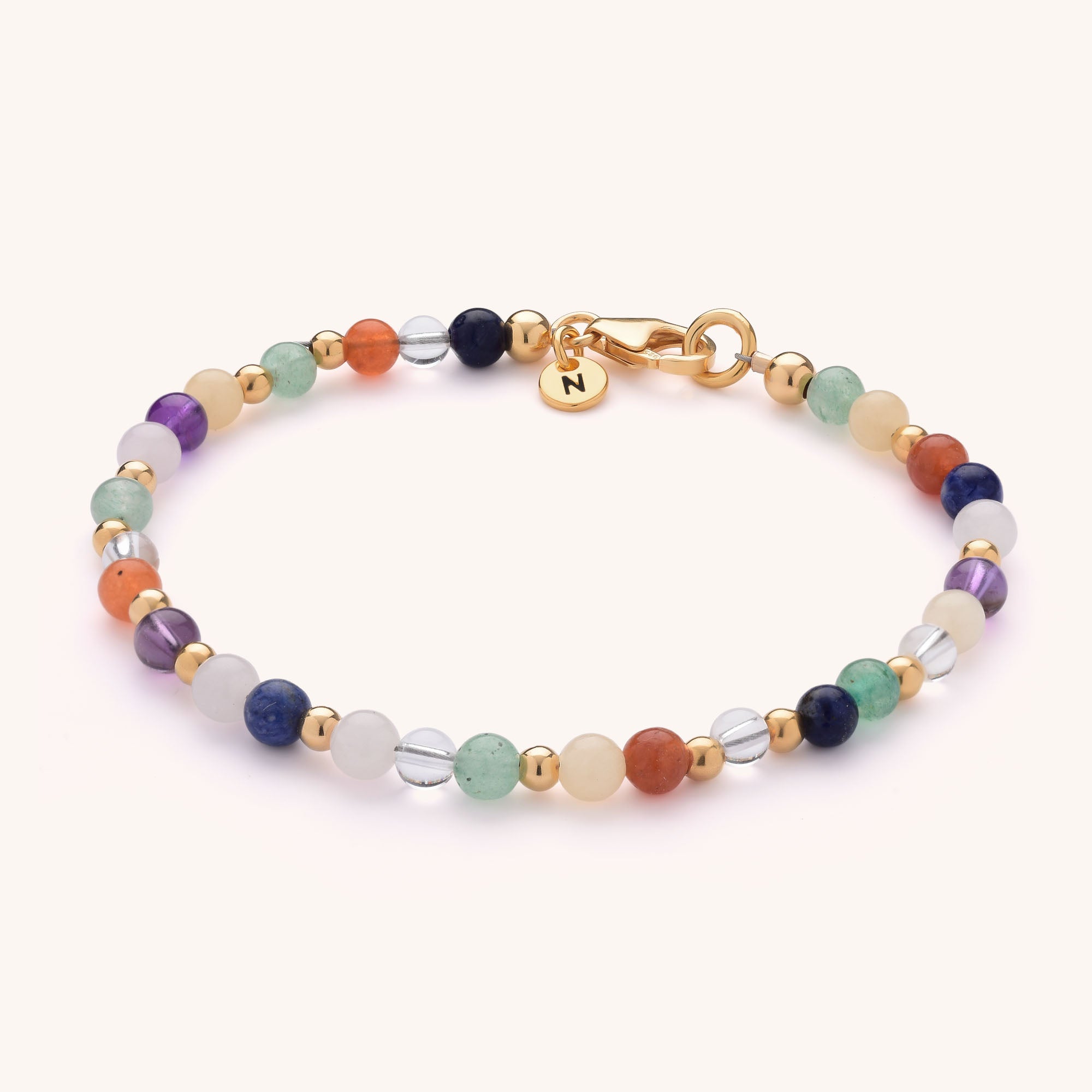 The Jewel Bead Chakra Bracelet features colorful natural gemstones, small gold beads, a gold clasp, and a round gold charm engraved with the letter N.