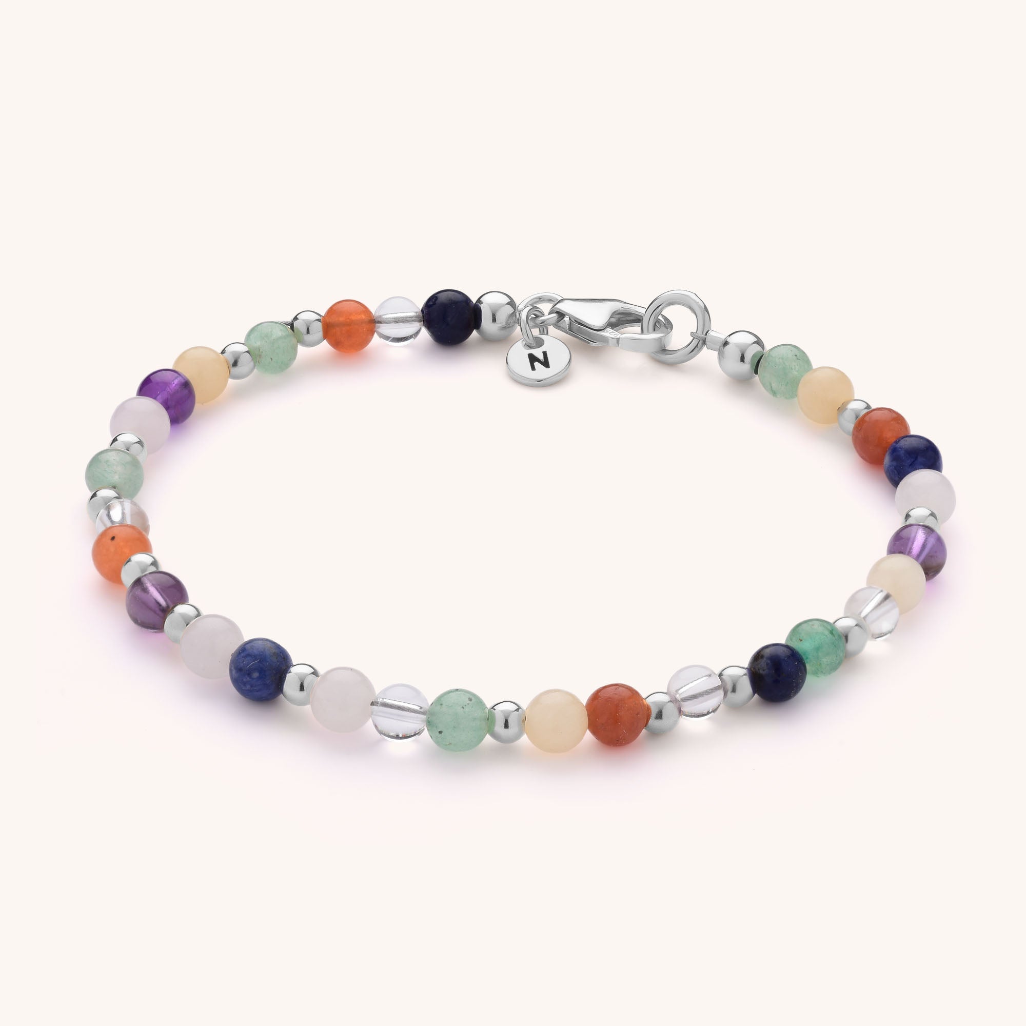The Jewel Bead Chakra Bracelet features multicolored natural gemstones, sterling silver beads, a silver clasp, and a small round tag engraved with the letter N.