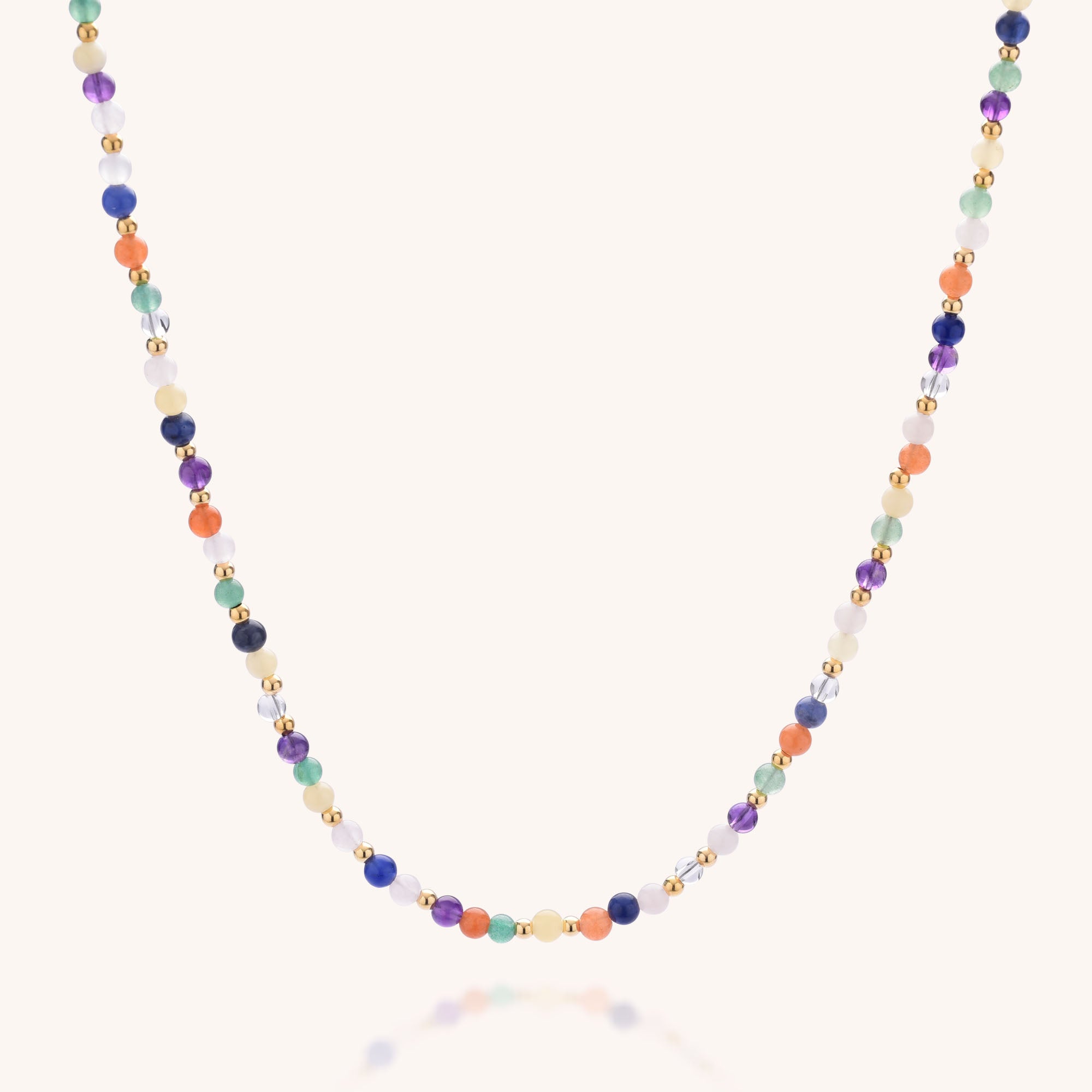 The Jewel Bead Chakra Necklace showcases small, round natural gemstones in blue, orange, green, purple, and gold, arranged symmetrically on a fine sterling silver chain against a light background.