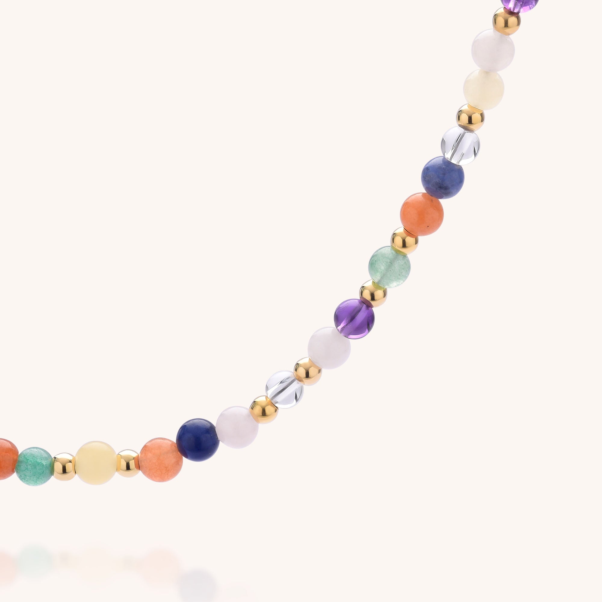 A close-up of the Jewel Bead Chakra Necklace, showcasing round, multicolored natural gemstones and small gold beads in a repeating pattern on a light background.