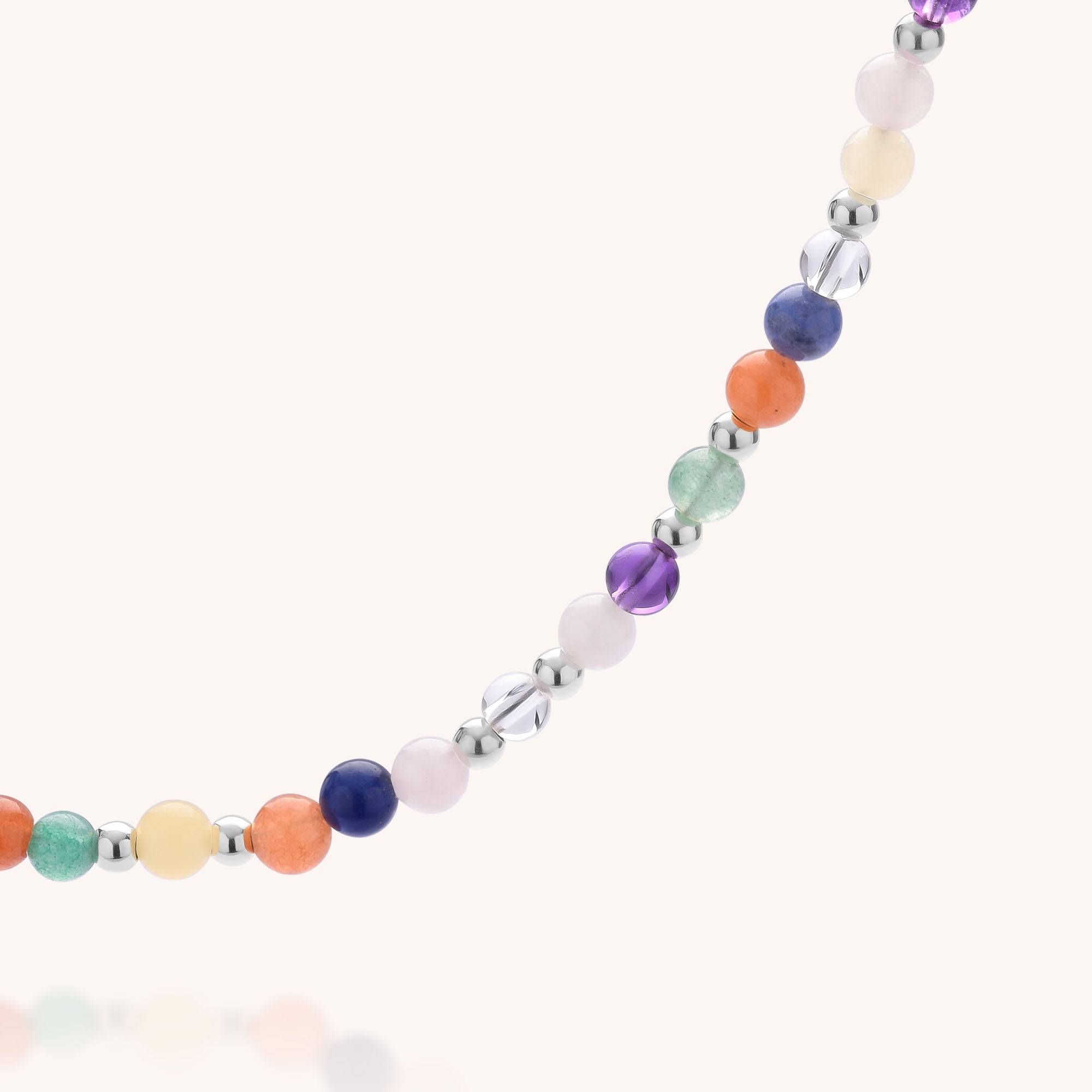 The Jewel Bead Chakra Necklace showcases round, colorful natural gemstone beads and small sterling silver spacers, elegantly arranged in a curved line against a light background.