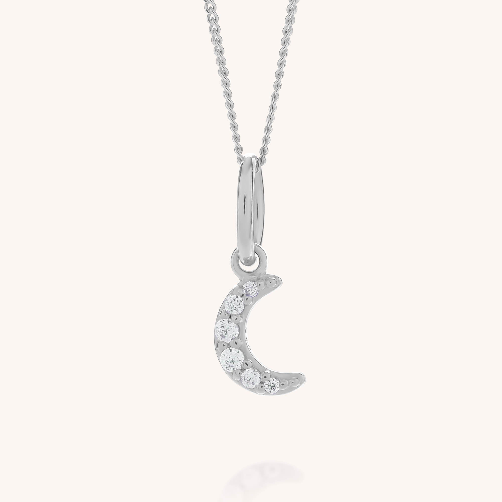 The Starlit Moon Necklace showcases a slender crescent moon pendant embellished with sparkling clear gemstones, skillfully crafted from eco-friendly sterling silver and set against a soft background.