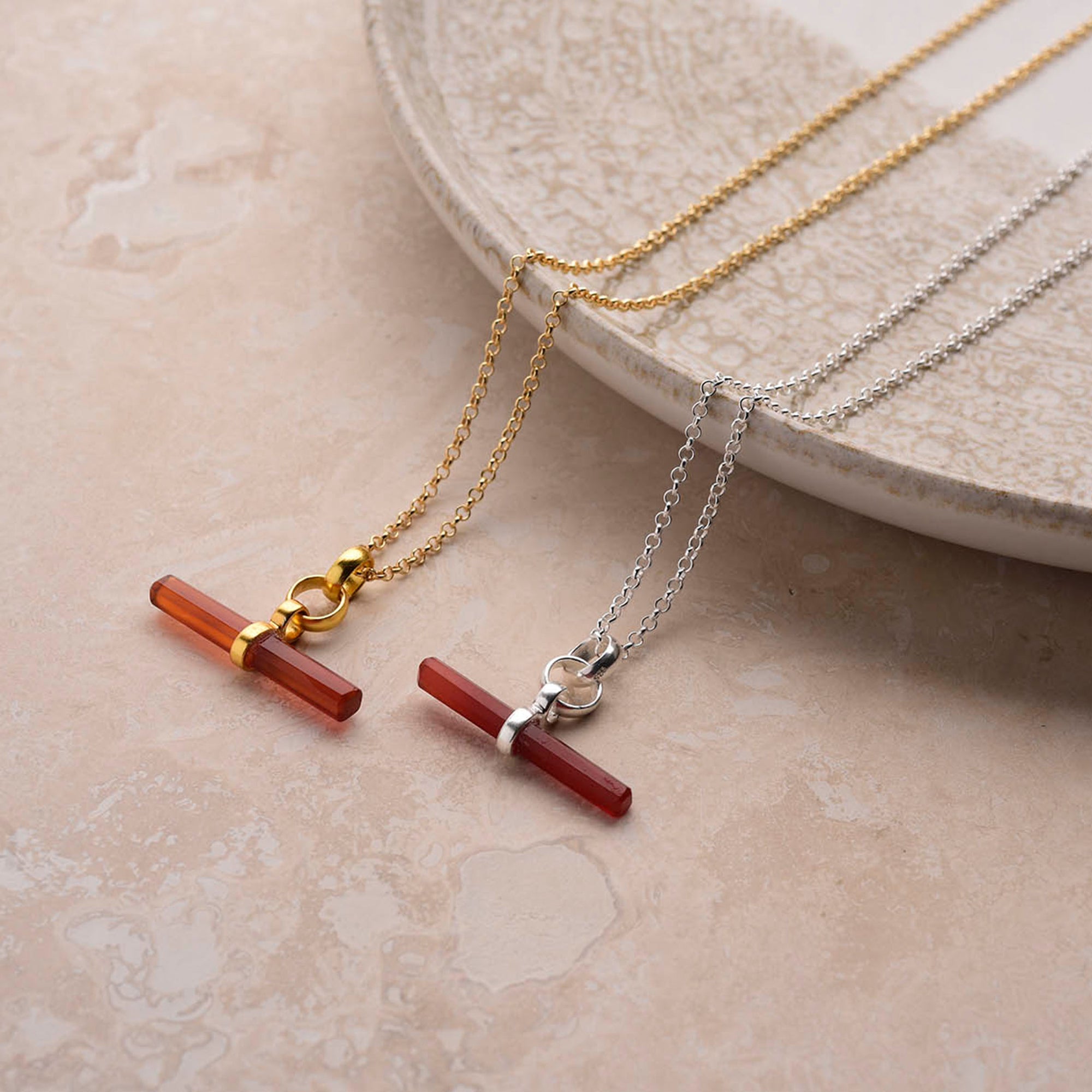 Two Gemstone T Bar Necklaces with horizontal red bar pendants rest on a beige surface near a ceramic plate. One has a gold chain, the other silver—each with matching metal accents on the pendant.