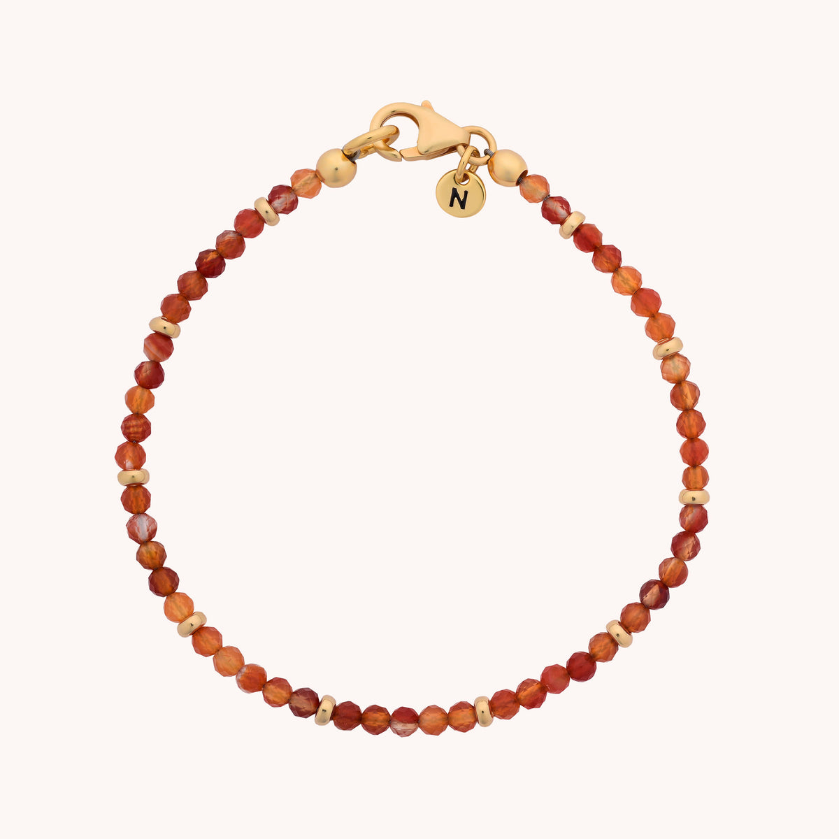 Introducing the Carnelian Orbit Bracelet - July: a handcrafted jewellery piece showcasing small, round carnelian beads in warm reddish-brown hues, complemented by gold spacer beads and finished with a gold clasp. A delicate, round gold charm engraved with the letter “N” adds a personal touch. The bracelet is elegantly arranged in a circle against a simple background.