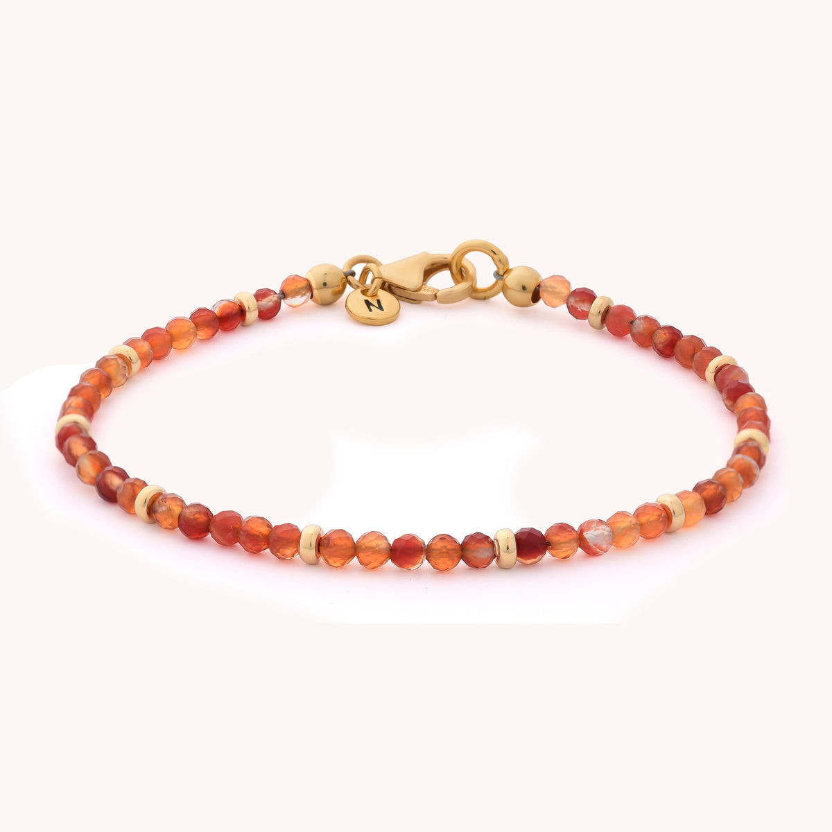 The Carnelian Orbit Bracelet - July is a delicate handcrafted jewellery piece featuring vibrant orange and beige beads reminiscent of carnelian, complemented by a gold clasp and a small round gold charm engraved with the letter N.